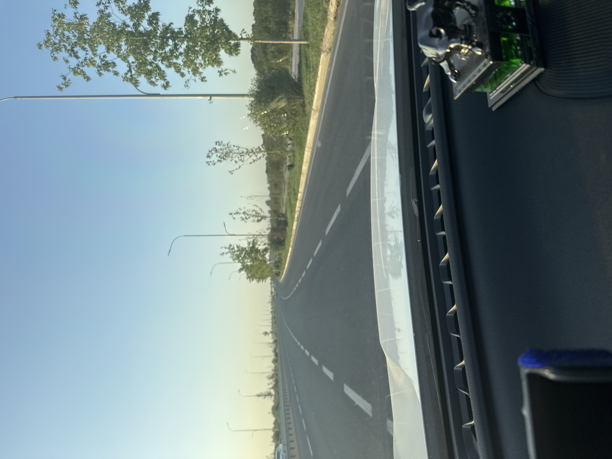 The image shows a view from inside a car, looking out at a road in Dahbet, Uzbekistan. The road is paved with asphalt and has white dashed lines marking the lanes. There are streetlights on both sides of the road, and trees and greenery line the roadside. The sky is clear and blue. In the foreground, on the car's dashboard, there is a small black bull figurine.
