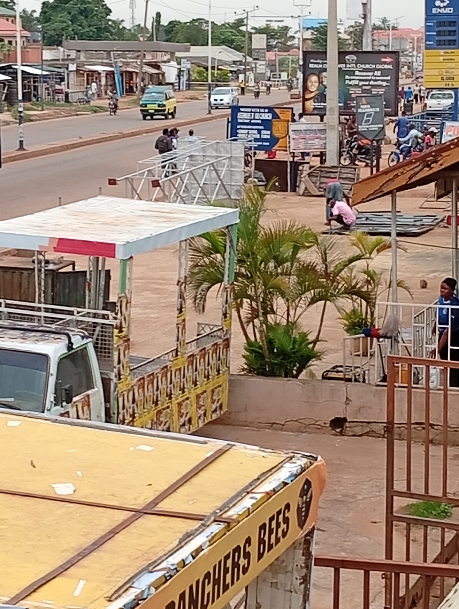 The image depicts a street scene in Sabon Tasha, Nigeria. A busy road with vehicular traffic is visible, including cars and motorcycles. Pedestrians are present on the roadside. A truck with a canopy is parked. In the foreground, there are partial views of buildings with signage, including one reading "RANCHERS BEES". Several individuals are seen engaging in various activities, including squatting and standing near a metal structure. There are plants, metal construction materials, and advertisements on billboards. A woman in a pink shirt is squatting. A woman wearing a blue top is standing near a gated area. The environment appears to be a mix of commercial and residential areas under overcast lighting.
