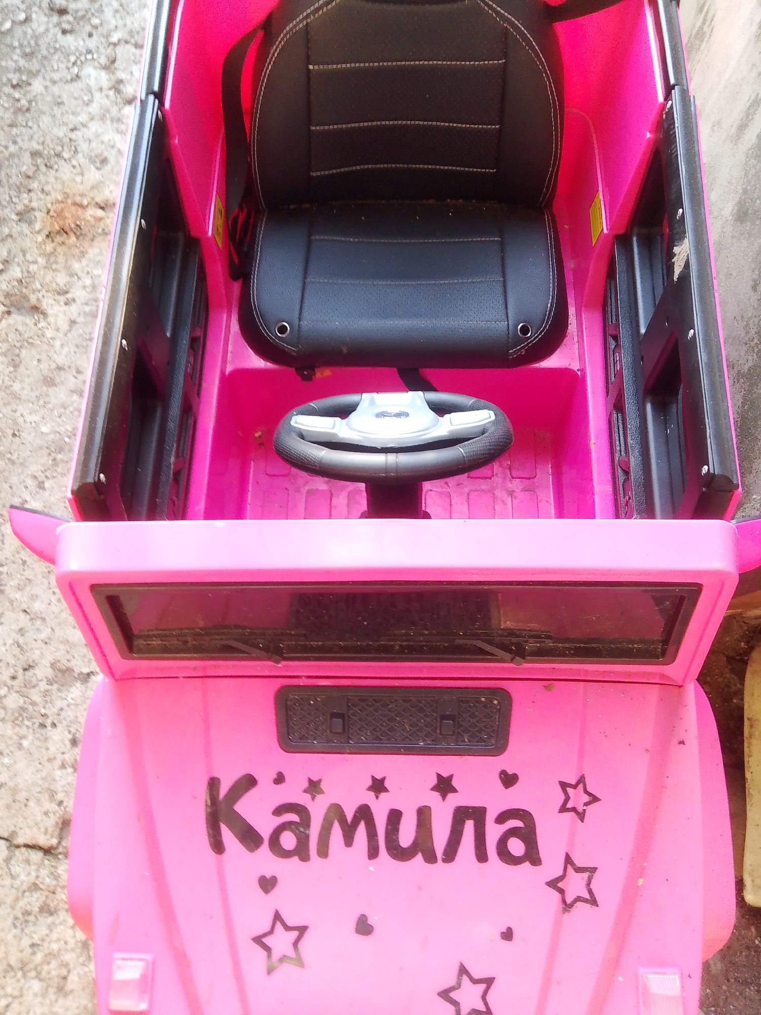 The image shows a pink toy car parked on a cracked concrete surface. The car has a black leather seat with white stitching, a black steering wheel, and the name "Камила" written in black letters on the front hood, surrounded by stars and hearts. The environment appears to be an outdoor setting, possibly a yard or street, in Хрестівка, Ukraine. The lighting suggests it is daytime. No people are present in the image.
