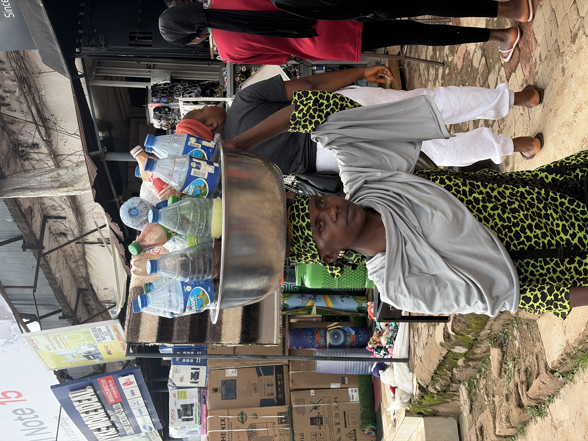 A street scene in Jos, Nigeria, features a woman carrying a large metal bowl filled with water bottles on her head. She wears a green and black leopard-print dress and a grey shawl. Behind her, another woman in a red shirt and black headscarf stands, and to her right, a person wearing grey sweatpants and a black headscarf is visible. In the background, various shops and stalls display goods, including electronics, appliances, and other merchandise. The ground is paved with bricks and dirt. The lighting suggests it is daytime, possibly overcast.
