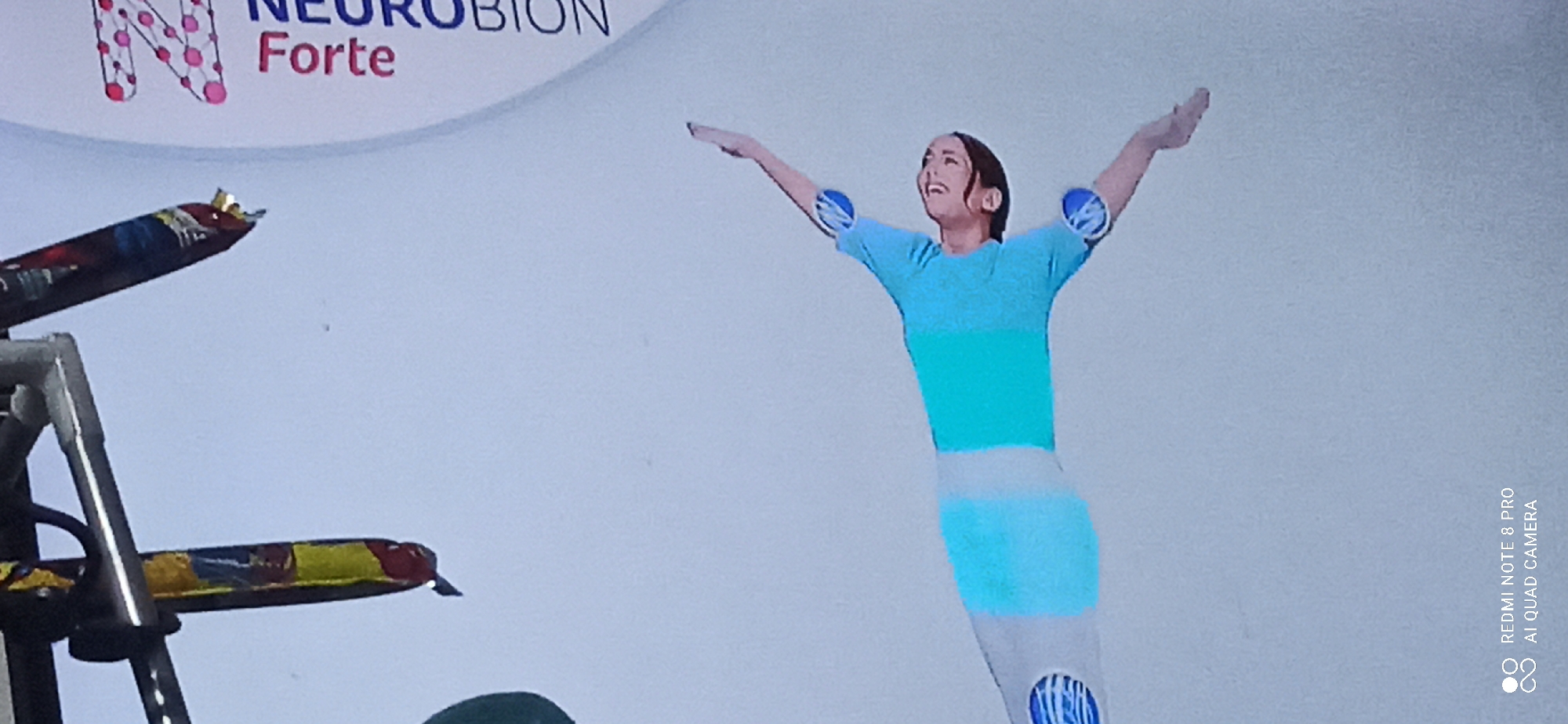 The image depicts a gymnast in a dynamic pose against a plain, light blue background. The gymnast is wearing a blue and white leotard with blue circular accents on the elbows. The gymnast's arms are extended upwards and outwards, and their head is tilted back, with an open-mouthed expression suggesting exertion or exhilaration. The background is featureless, with the exception of some text in the upper right corner which reads "Neurobion Forte". In the upper left corner, a blurry object with colorful bands, possibly packaging or a flag, hangs down. The bottom right corner of the image contains text that reads "REDMI NOTE 8 PRO AI QUAD CAMERA", indicating the device used to capture the photograph. The image is oriented vertically, with the gymnast appearing to be in mid-air. There is no discernible urban or natural environment, therefore the location in Sedati Agung, Indonesia, cannot be determined from the visual information provided.
