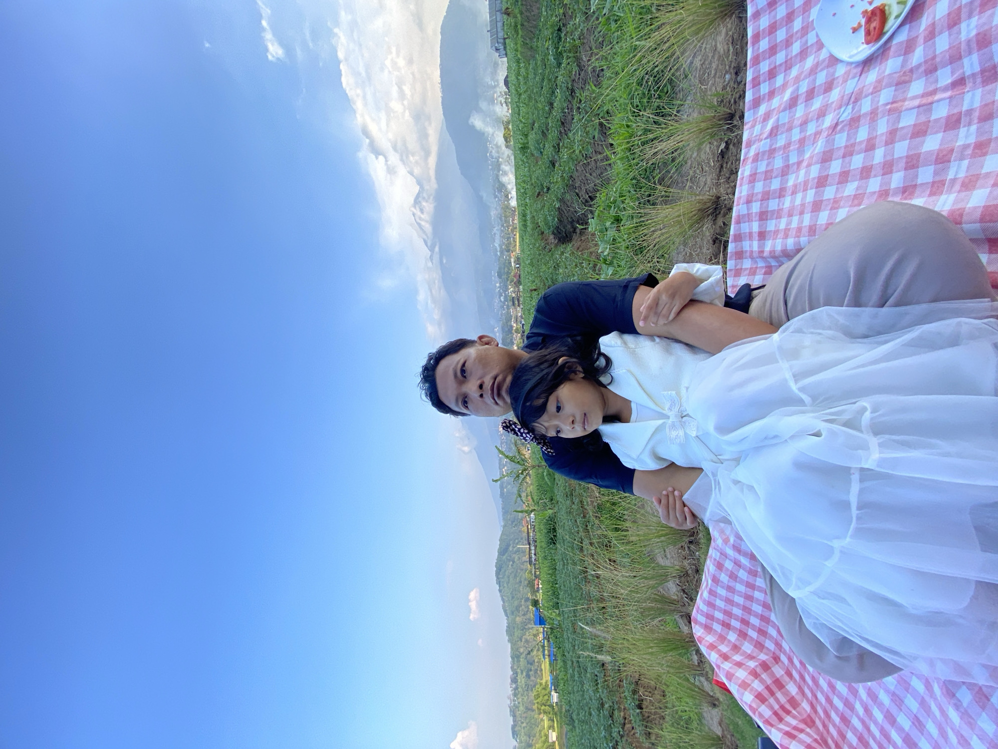 An adult male and a young girl are seated on a red and white checkered blanket in a field. The male, wearing a dark blue shirt, has his arm around the girl. The girl, dressed in a white dress, is seated on his lap. The background features a lush green field with rows of crops, mountains in the distance, and a clear blue sky. The scene suggests a picnic or outdoor gathering. The location is the city of Kesiman, Indonesia.