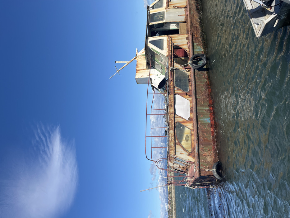 An old, rusted boat sits in the water on a clear day. The boat is tall with multiple levels and rectangular windows. Rust covers most of the boat's exterior, and tires are attached to the side. A railing is visible along one edge. The water is rippled, and a blue sky with a single, prominent cloud is in the background.