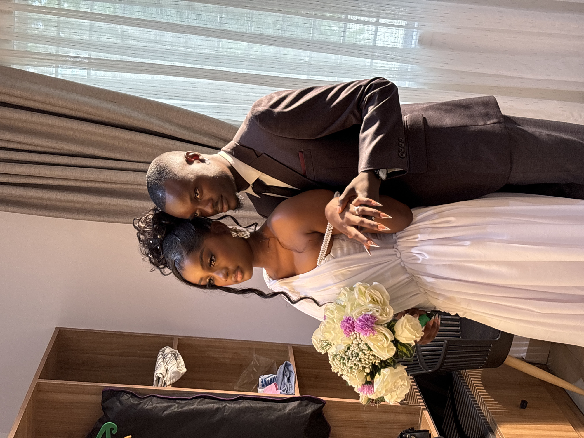 A newlywed couple is posing for a photograph. The man is wearing a brown suit, white shirt, and striped tie. The woman is wearing a white dress and holding a bouquet of white and purple flowers. They are standing indoors next to a window with curtains. In the background, there is a wooden cabinet. The location is presumably Gura Top, Nigeria, based on the prompt's request.