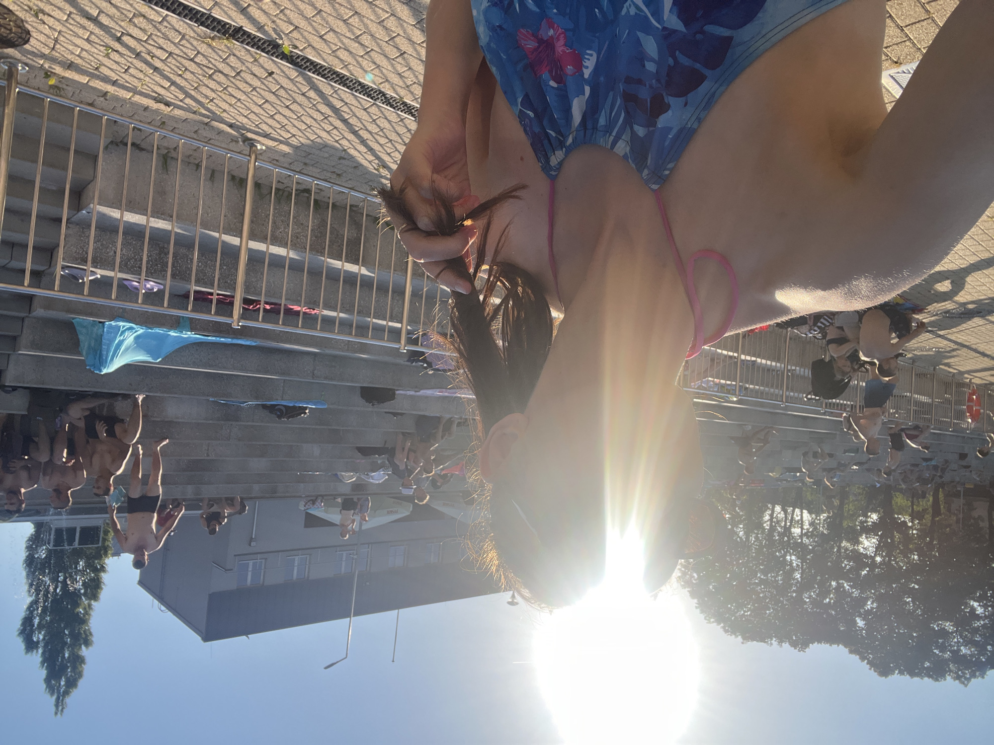 This is a medium shot of a group of people on steps near a body of water, likely a swimming pool or a lake, on a sunny day. The perspective is skewed, with the subjects appearing sideways. The main subject in the foreground is a person wearing a blue and white patterned swimsuit, pulling on their dark hair. They are positioned at the top of the frame, and the bright sun is shining directly into the lens, creating a strong lens flare that obscures some details.
In the background, on a tiered set of concrete steps, are several other individuals, mostly men, some wearing swim trunks. They appear to be relaxing or possibly preparing to enter the water. A metal railing runs along the top of the steps. A blue towel is draped over the railing in the mid-ground. Beyond the steps, a modern-looking building with multiple windows is visible. To the left, a tall, leafy tree stands out against the clear blue sky. The bright sunlight suggests it is daytime, likely midday or afternoon, and the weather is warm and clear. The overall mood appears casual and summery, with people enjoying an outdoor recreational space. No visible text is present in the image.