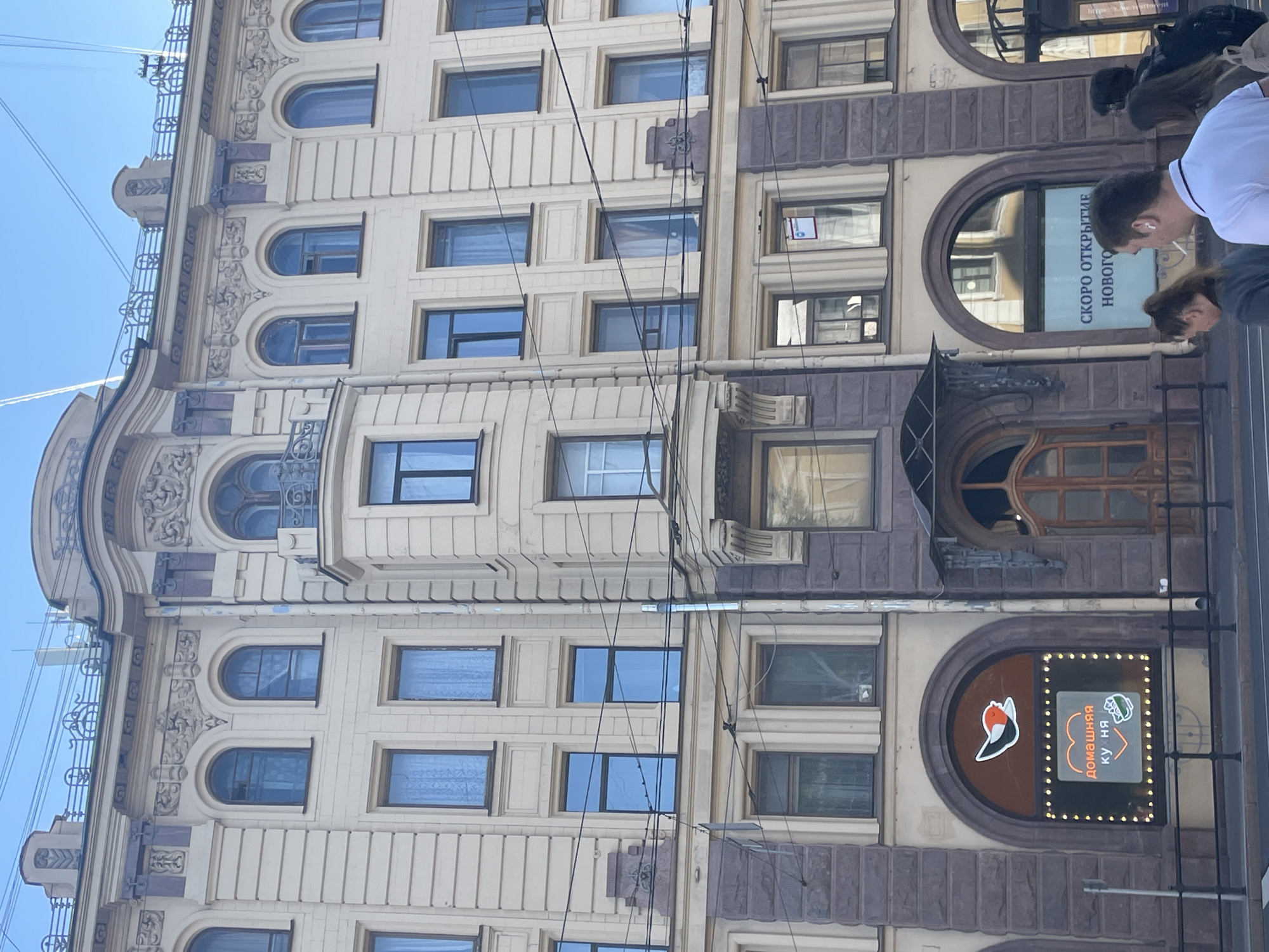 The image shows a multi-story building facade in Saint Petersburg, Russia. The building has several floors with numerous windows of varying shapes and sizes, many of which are arched. The facade is decorated with architectural details, including ornate carvings and cornices. On the ground floor, there are two arched entrances with shops. One shop has a sign with a bird graphic and the text "ДОМАШНЯЯ КУХНЯ" (Homemade Cuisine) in Russian, illuminated by a string of lights. Next to it, another shop has a sign with Russian text that appears to say "СКОРО ОТКРЫТИЕ НОВОГО" (Soon to open new). Several people are walking on the sidewalk in front of the building. Overhead, there are telephone wires and a clear blue sky with faint contrails. The architecture suggests an older, classic European style typical of Saint Petersburg.