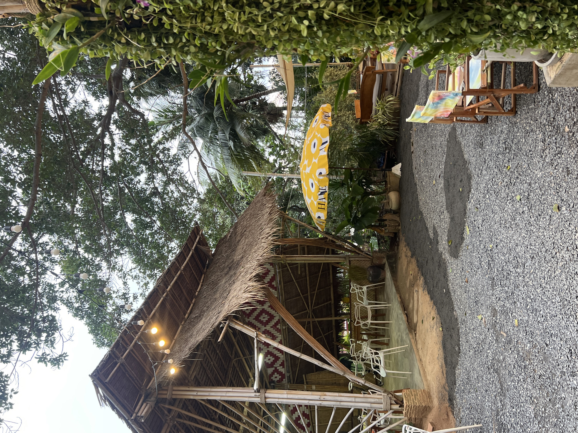 This media file depicts an outdoor seating area at a cafe or restaurant in Karon, Thailand. The scene is set with a traditional thatched-roof structure, supported by bamboo poles, providing shade. String lights are visible hanging above the structure, suggesting it might be used in the evening. White plastic chairs are arranged at tables within the covered area.
Outside the thatched structure, a gravel path leads to other seating arrangements. These include wooden chairs and tables, some with colorful cushions, and lounge chairs with tie-dye style fabric. A prominent yellow umbrella with a floral pattern is open, providing shade. Some greenery, including palm fronds and vines, is visible throughout the scene, indicating a tropical environment.
The lighting suggests it is daytime, possibly overcast, as there are no harsh shadows. The overall atmosphere appears relaxed and inviting, suitable for a casual dining or relaxation spot. Text is visible on the yellow umbrella, though it is partially obscured and difficult to read clearly. The word "HAVE" is discernible, possibly part of a longer phrase.