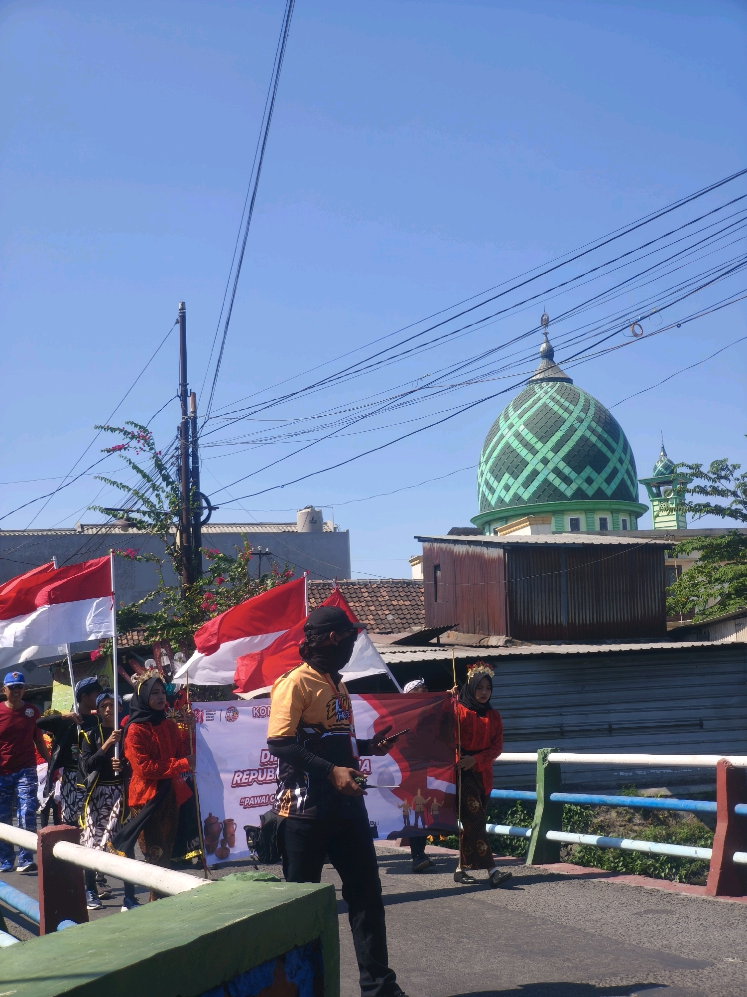 The image captures a sunny outdoor scene in Kepuh Kiriman, Indonesia, with a procession taking place. In the background, a distinctive mosque with a green, patterned dome and a smaller minaret is visible. The sky is clear blue, with power lines crossing horizontally across the frame. In the foreground, several people are walking, some carrying Indonesian flags (red and white). One woman in the center is dressed in a vibrant orange outfit and a headscarf, holding a flag. To her right, a man wearing an orange and black shirt with a black mask covering his lower face is walking. A banner with Indonesian text and illustrations is also visible, suggesting a patriotic or celebratory event. The procession appears to be on a street with a bridge or elevated walkway to the right, bordered by a railing. The overall atmosphere is lively and suggests a community gathering.