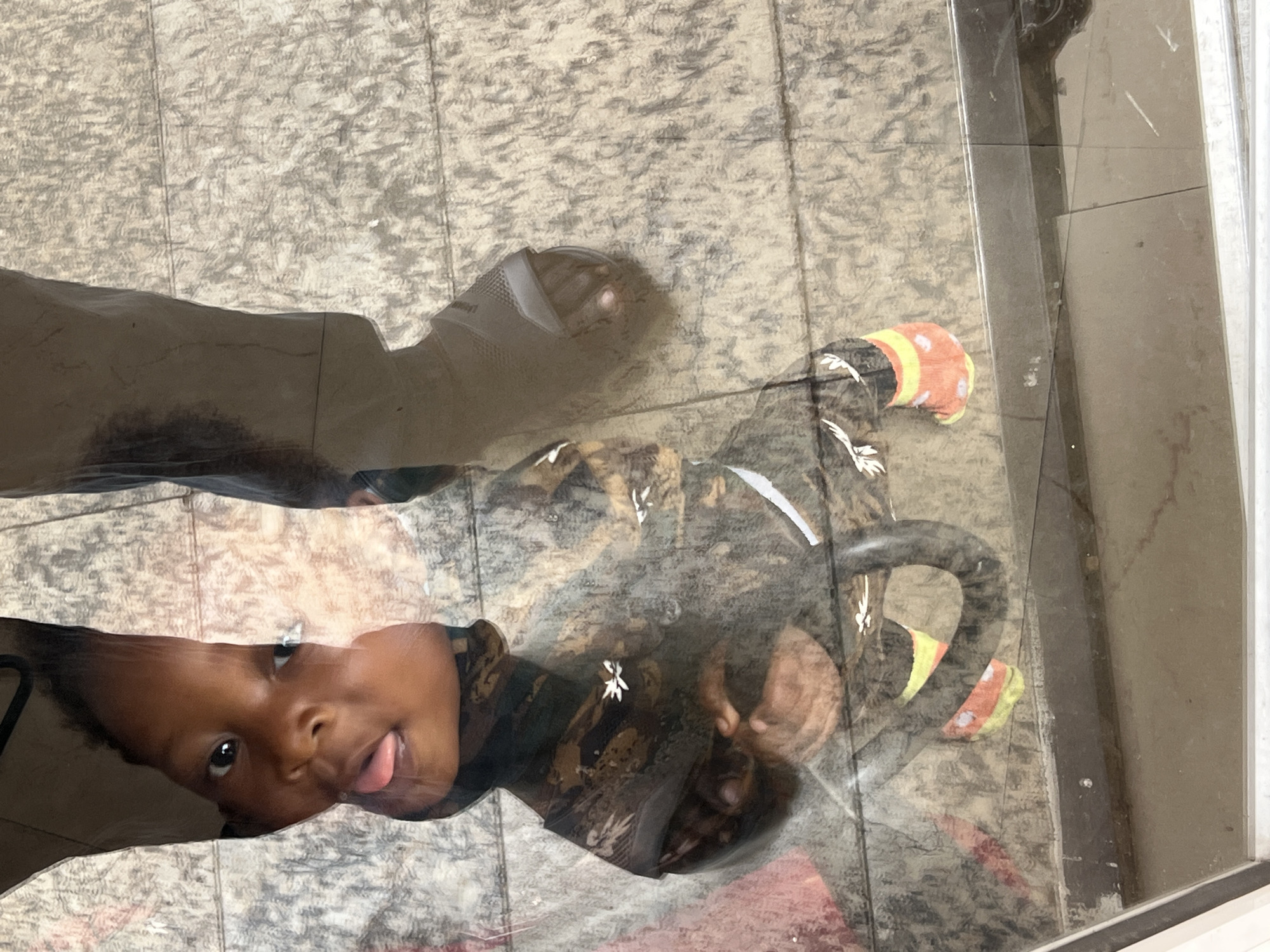 A young, dark-skinned child lies prone on a surface, possibly glass. The child sticks their tongue out. An adult figure is partially visible above the child. The reflection on the glass surface creates a superimposed image of the child and the adult's shadow against a flooring pattern. The location is not identifiable as Gura Top, Nigeria.