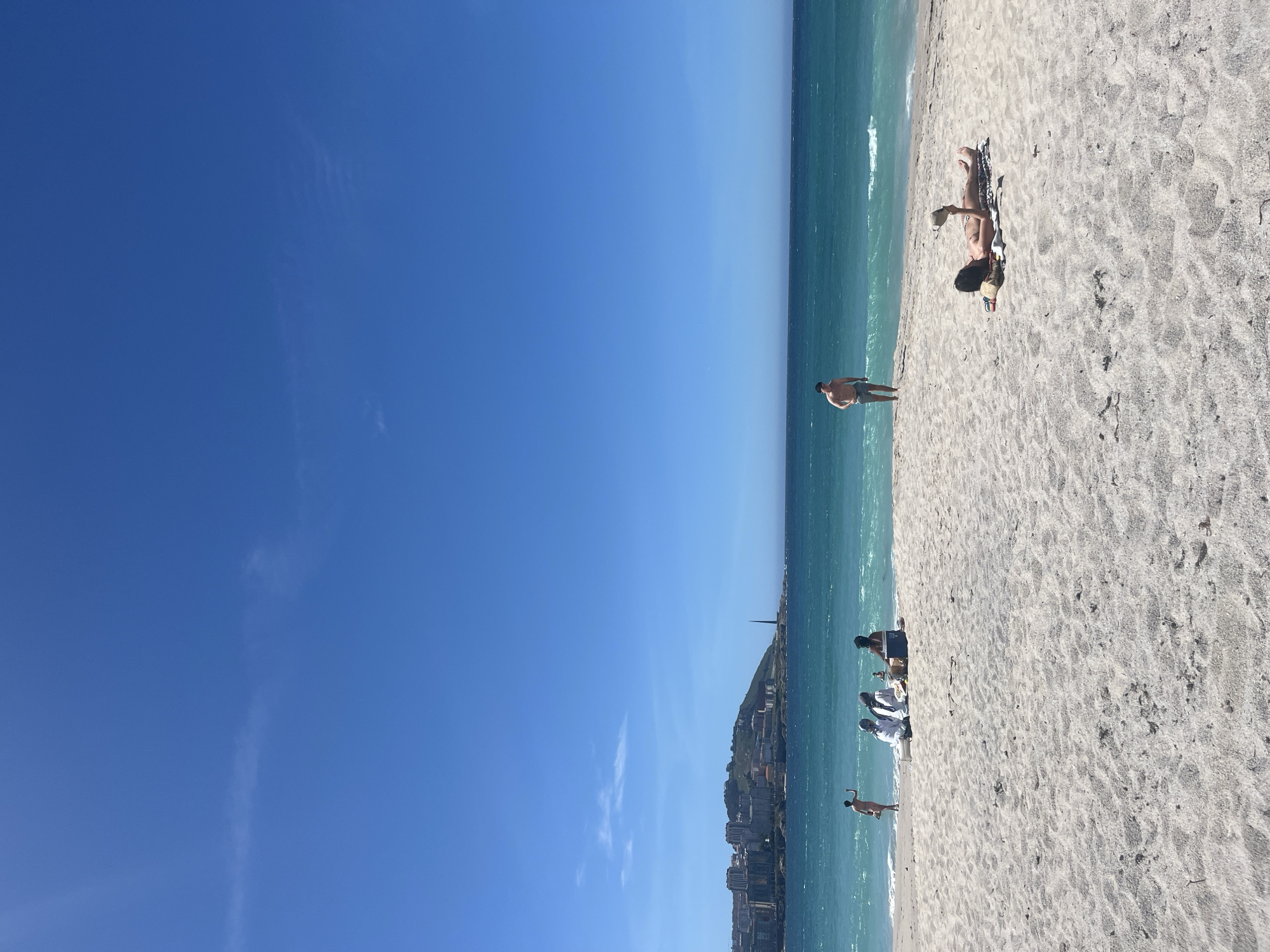 The image shows a scenic view of a sandy beach on a sunny day. The sky is clear and blue with a few wisps of clouds. The ocean water is a vibrant turquoise, with gentle waves lapping at the shore. In the distance, a city skyline with buildings and a distinctive tower rises on a hill.
On the beach, there are several people. Towards the right, a person is lying on a towel, appearing to be sunbathing and holding a hat. Further left, a group of people are sitting on the sand, possibly having a picnic. In the water, a person is standing near the shore, and another person is wading or swimming. A man in swimming trunks is standing at the edge of the water on the left side of the frame. The sand is light-colored and textured.
The lighthouse visible in the distance is likely the Tower of Hercules, a UNESCO World Heritage site and the oldest Roman lighthouse in the world, which is located in the city of A Coruña, Spain. The city appears to be situated on a peninsula jutting into the sea.