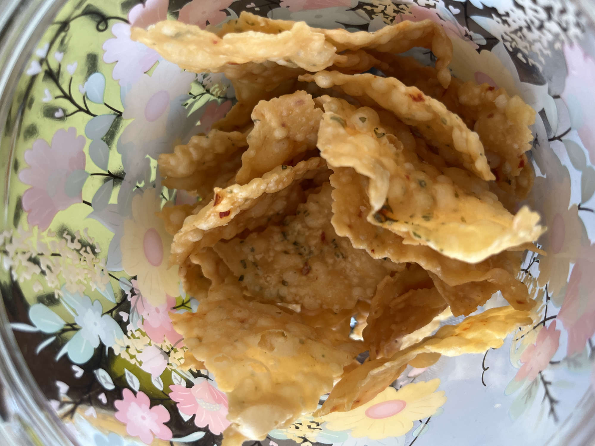 An elevated, close-up shot from within a clear container reveals a pile of irregularly shaped, fried dough chips. These chips are golden-yellow, with a bubbled texture and what appear to be herbs and possibly chili flakes scattered on their surfaces. The container has a floral pattern, featuring pastel shades of pink, yellow, and blue flowers on a light green background. The image suggests a food item, possibly a snack, within a decorative container. There is no visible information that connects the image to Lhokseumawe, Indonesia.