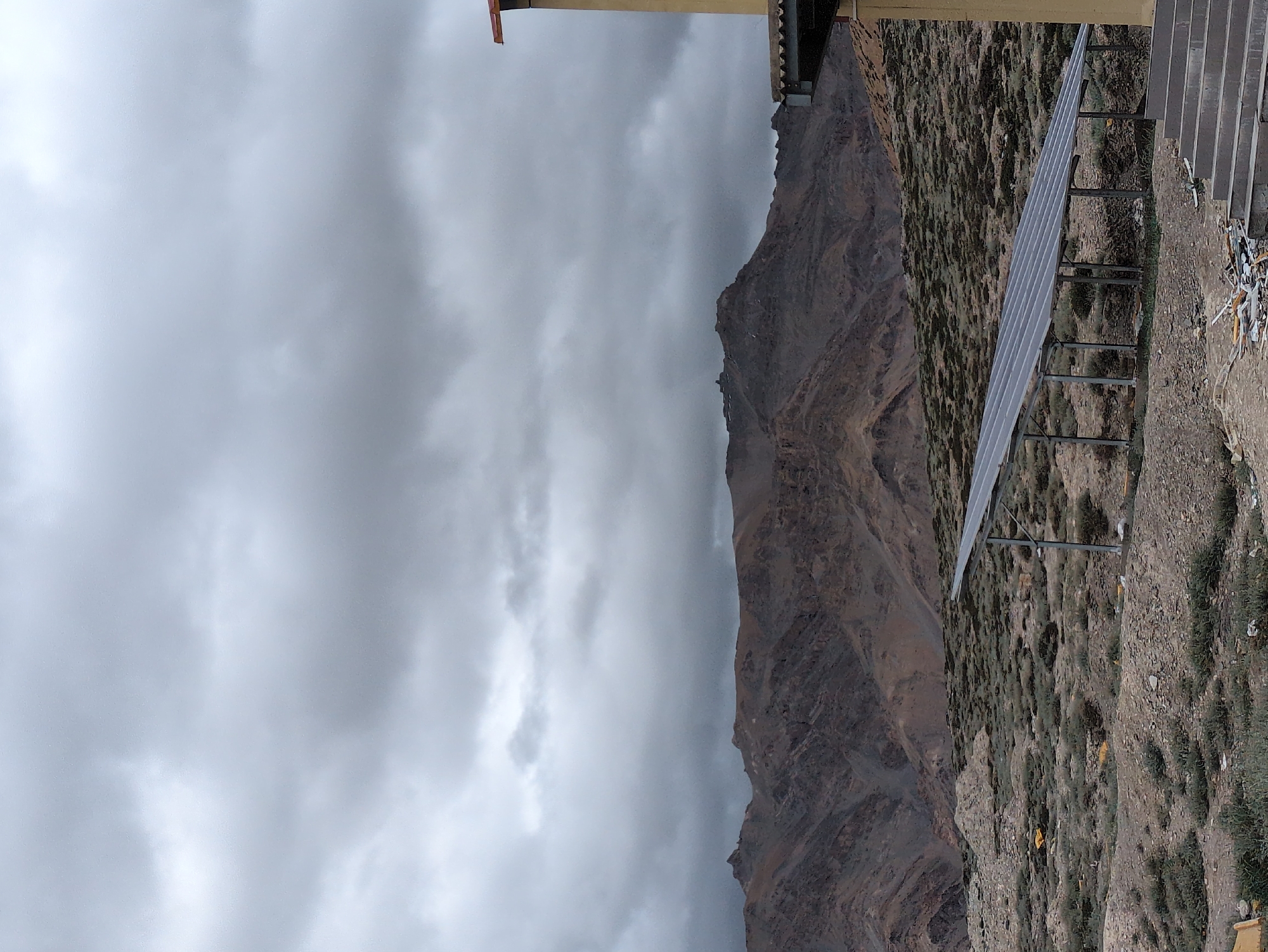 A solar panel array is positioned in the foreground of a rugged, mountainous landscape under a cloudy sky. The mountains are barren and appear to be made of rock and soil, with layered geological formations visible. Sparse vegetation, low-lying shrubs and grasses, dots the ground around the solar panels. To the right, a portion of a building with a yellow facade and a concrete staircase leading up to it is visible. The sky is filled with heavy, gray clouds, suggesting overcast weather. The overall scene depicts a somewhat remote, possibly arid, environment where renewable energy infrastructure has been installed. There are no people or animals visible in the image. The location, Dhar Chharp Samba, India, suggests a high-altitude, potentially mountainous region.