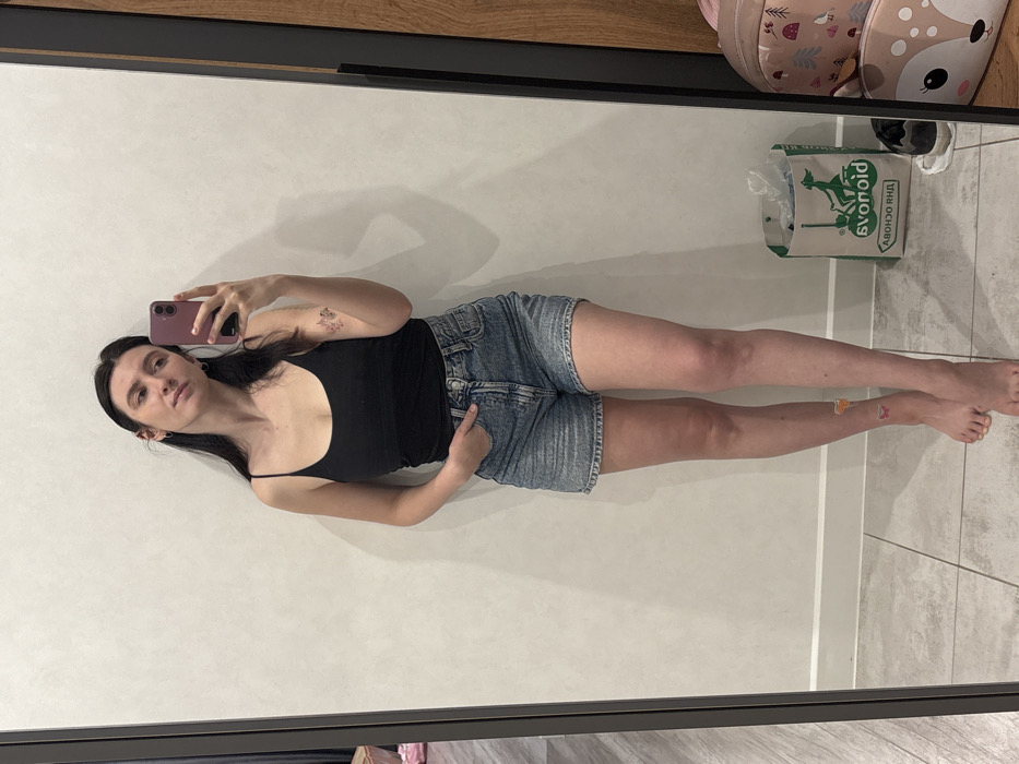 A young woman poses for a mirror selfie. She wears a black tank top and denim shorts, her left hand tucked in her pocket, and holds a rose-colored phone with her right hand. The mirror reflects a glimpse of a backpack with an animal print in the top right corner. On the floor, a shopping bag stands next to discarded shoes. A small butterfly sticker adorns her lower leg.