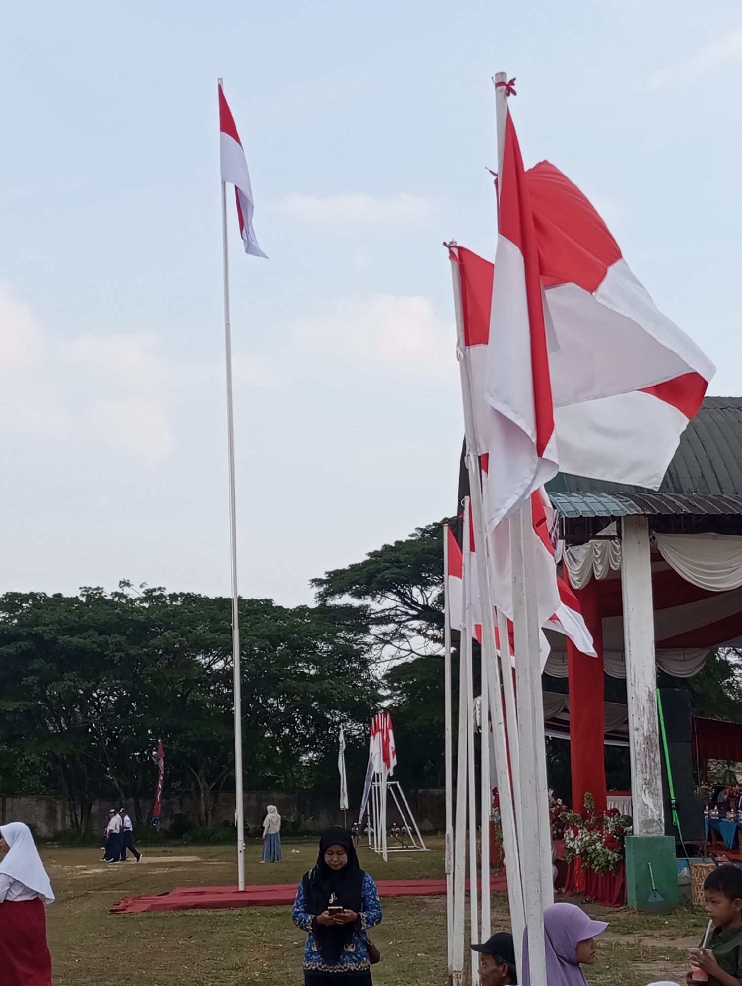 A low-angle, full shot captures an outdoor scene with Indonesian flags prominently displayed against a pale blue sky. In the foreground, a row of white flagpoles stand vertically, each adorned with red and white Indonesian flags that are billowing in the wind. The flags are positioned to the right of center. Behind the flagpoles, a building with a dark roof and red and white striped awnings is visible. To the left of the flagpoles, a dense line of dark green trees forms a backdrop, with some lighter green foliage interspersed. In the far left background, two young people in school uniforms are walking across a grassy field. In the mid-ground, a woman in a black hijab and a blue and white patterned top is looking down at her phone. To her right, a young boy with a straw in a cup is visible. The ground is covered in a dry, greenish-brown grass. The overall impression is that of a public event or ceremony taking place in Candirejo, Indonesia.