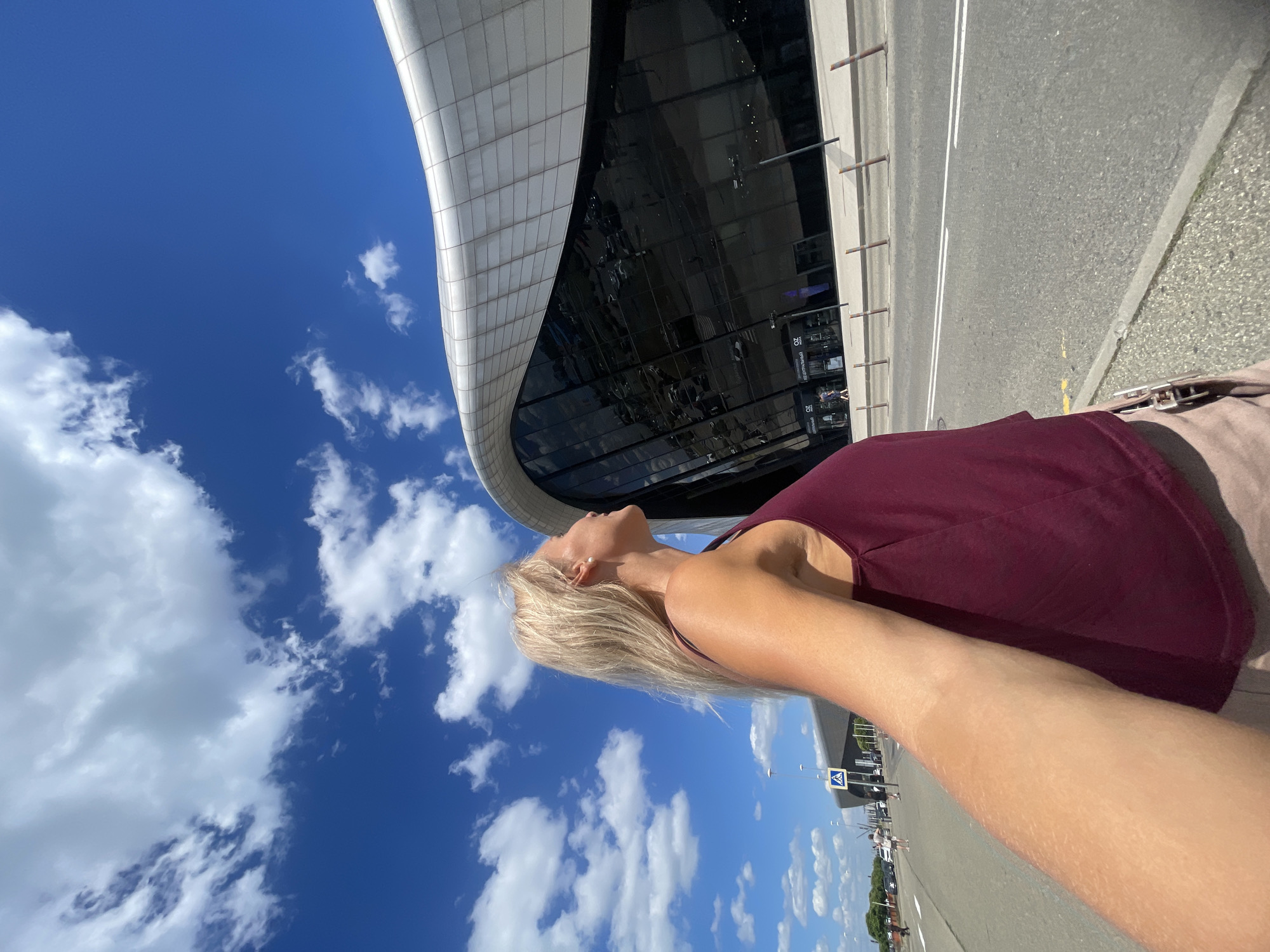 This is a medium-angle, slightly tilted, sunlit, outdoor, selfie-style photograph taken in Krasnodar, Russia, on a clear day with scattered white clouds. In the foreground and left side of the image, a woman with blonde hair pulled back, wearing a maroon tank top and light brown shorts, is captured from the chest up. She is looking upwards to her right, away from the camera, with her right arm extended, holding the camera.
In the background, a modern, curved, glass-fronted building with a silver, patterned facade dominates the scene. The building appears to be an entertainment or shopping complex, as indicated by signage for "OZ" and "OZ CENTRAL" visible on the entrance. A road with double white lines runs horizontally across the middle ground, with a sidewalk and a row of bollards separating it from the building. To the left of the building, there is a pedestrian crossing sign and several cars parked along the road. The overall environment suggests an urban setting, likely near a transportation hub or a public gathering space.