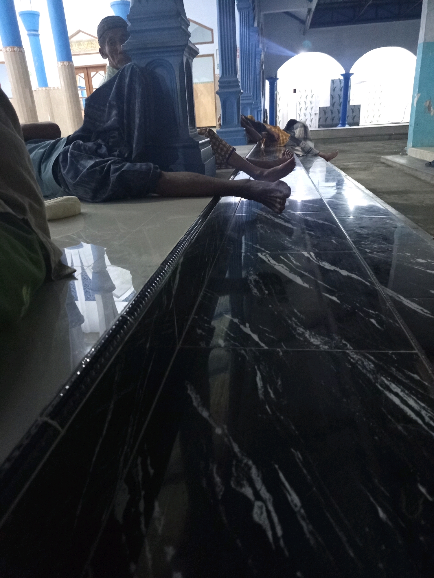 The image depicts the interior of a building, likely a mosque or a similar place of worship, in Ngablak, Indonesia. The foreground is dominated by a shiny, dark, marble-like tiled surface reflecting the surroundings. In the mid-ground, several individuals are seen resting or sleeping on the floor. One person, wearing a cap and a dark patterned garment, is reclined on their side, facing away from the camera, with their legs extended. Another individual is lying on their stomach in the background, partially covered by a patterned cloth. The architecture features tall, blue-colored pillars and arched doorways in the distance. The overall impression is one of a quiet, perhaps communal space where people are taking a rest.