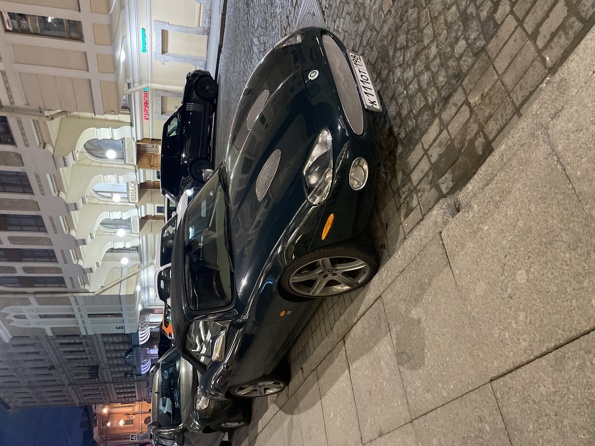 The image depicts a dark green Jaguar XK parked on a cobblestone street in Saint Petersburg, Russia. A black car is parked behind the Jaguar. The license plate on the Jaguar reads "K 110 OT 198". Buildings line the other side of the street, with architectural details including arches, windows, and street lights. Street signage is visible. The photo was taken at night.