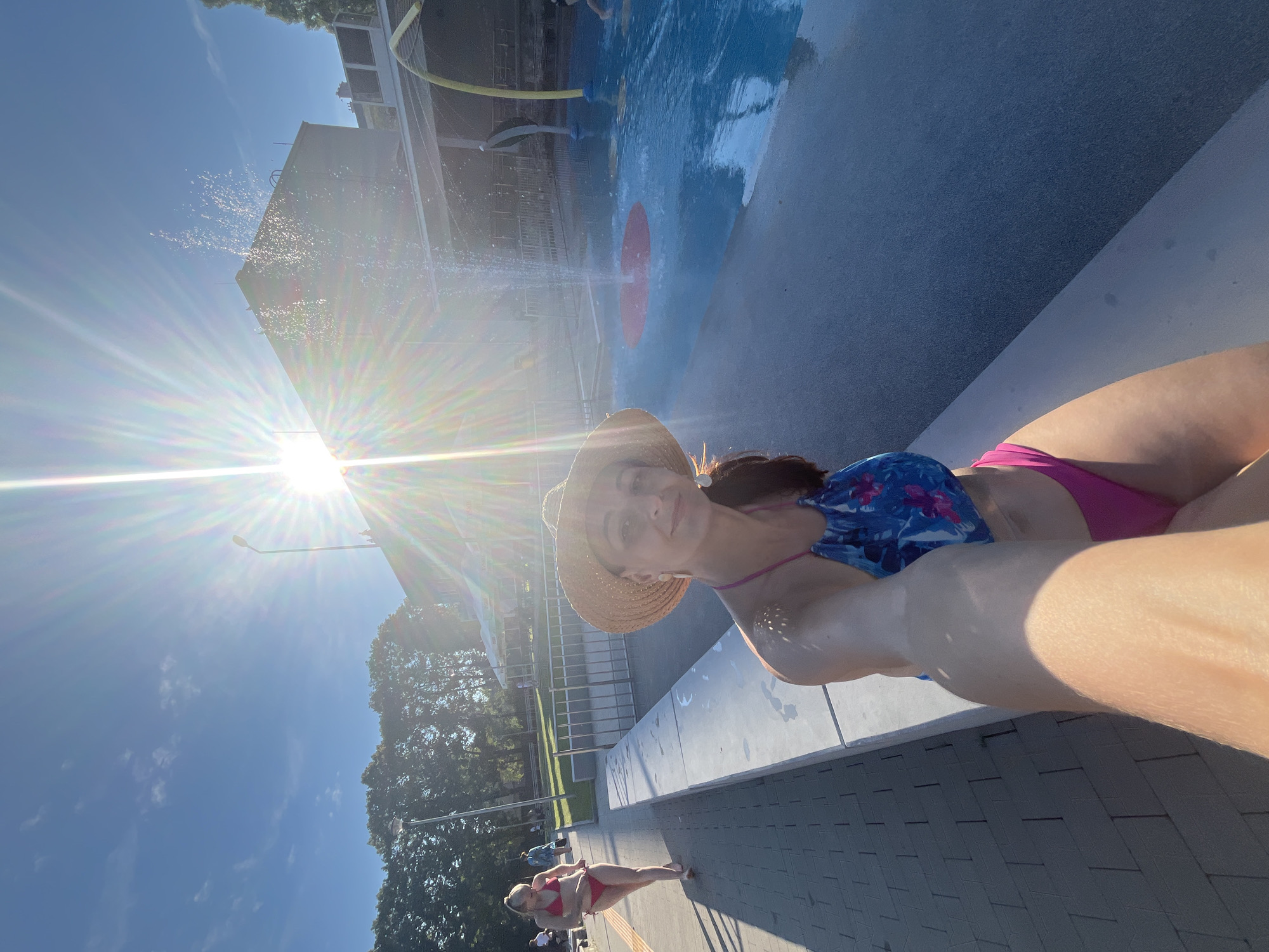 A person wearing a straw hat and a floral bikini is taking a selfie in a park with a splash pad. The sun is shining brightly, creating lens flare and casting shadows. In the background, water sprays from a structure, and a blue splash pad with a red circle is visible. Another person in a pink bikini is also visible in the background, lying on the ground. The scene appears to be outdoors on a sunny day, likely during warmer months. The overall mood seems relaxed and recreational.