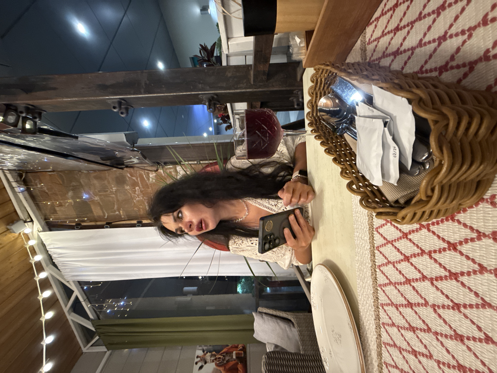 A woman with dark hair, wearing a white lace top and a pearl necklace, sits at a table in a restaurant. She is holding a black smartphone in her left hand and looking upwards with her mouth open. In front of her is a glass of red wine and a white plate. A basket containing cutlery and paper packets is on the table. The restaurant appears to be an outdoor seating area with a wooden ceiling and brick walls. String lights are visible overhead. A framed picture of a giraffe wearing a suit is on the wall in the background. The image is taken at night. This scene is in Sochi, Russia.
