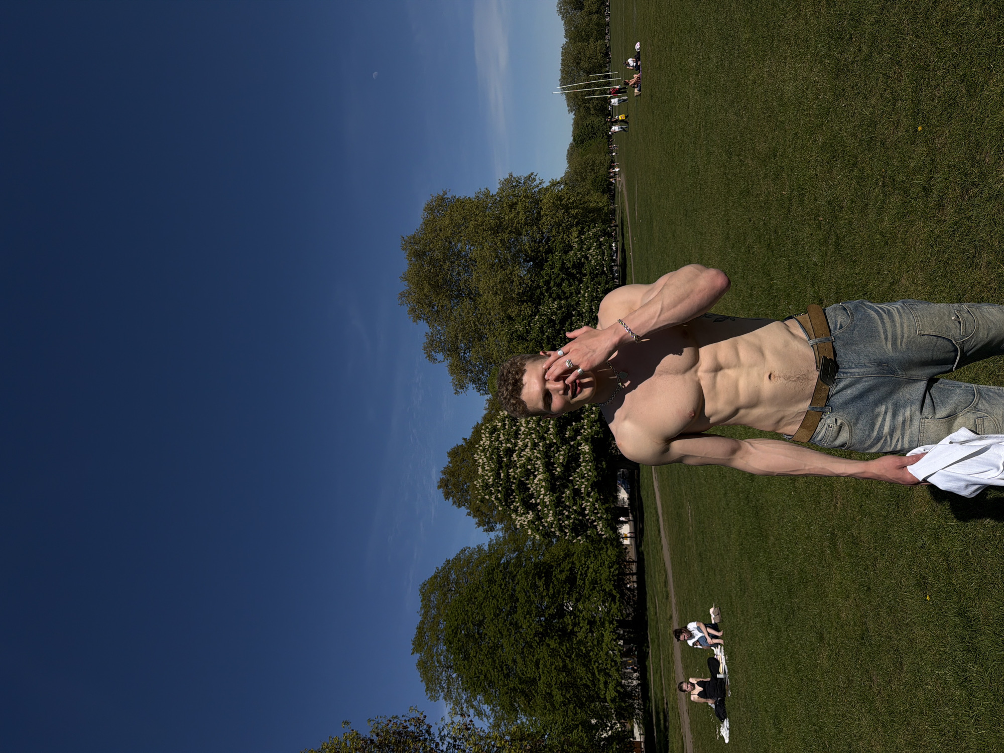A shirtless man stands in a grassy park, wearing jeans and shielding his eyes with his hand. The sky is bright blue. Other people relax on the grass behind him. The location appears to be a park in London, United Kingdom.