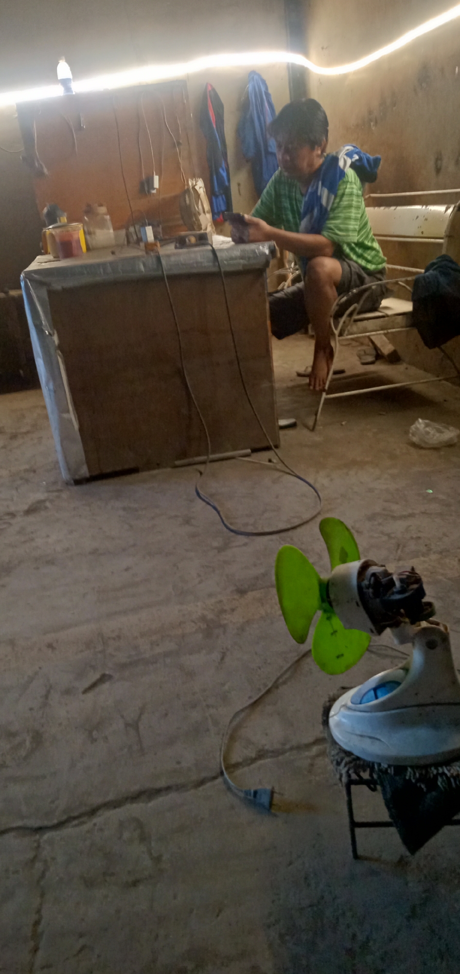 The image depicts an indoor scene in Sumput, Indonesia. A person wearing a green striped shirt and dark shorts sits at a wooden table and appears to be using a mobile phone. A blue and white towel rests over their shoulders. The table is cluttered with various items and covered with a plastic sheet. A small, white and green electric fan sits on a black metal stand in the foreground. Wires are visible, running from the table to the wall and to the fan. The room has a concrete floor and appears to be a workspace, with clothes hanging on the wall in the background.