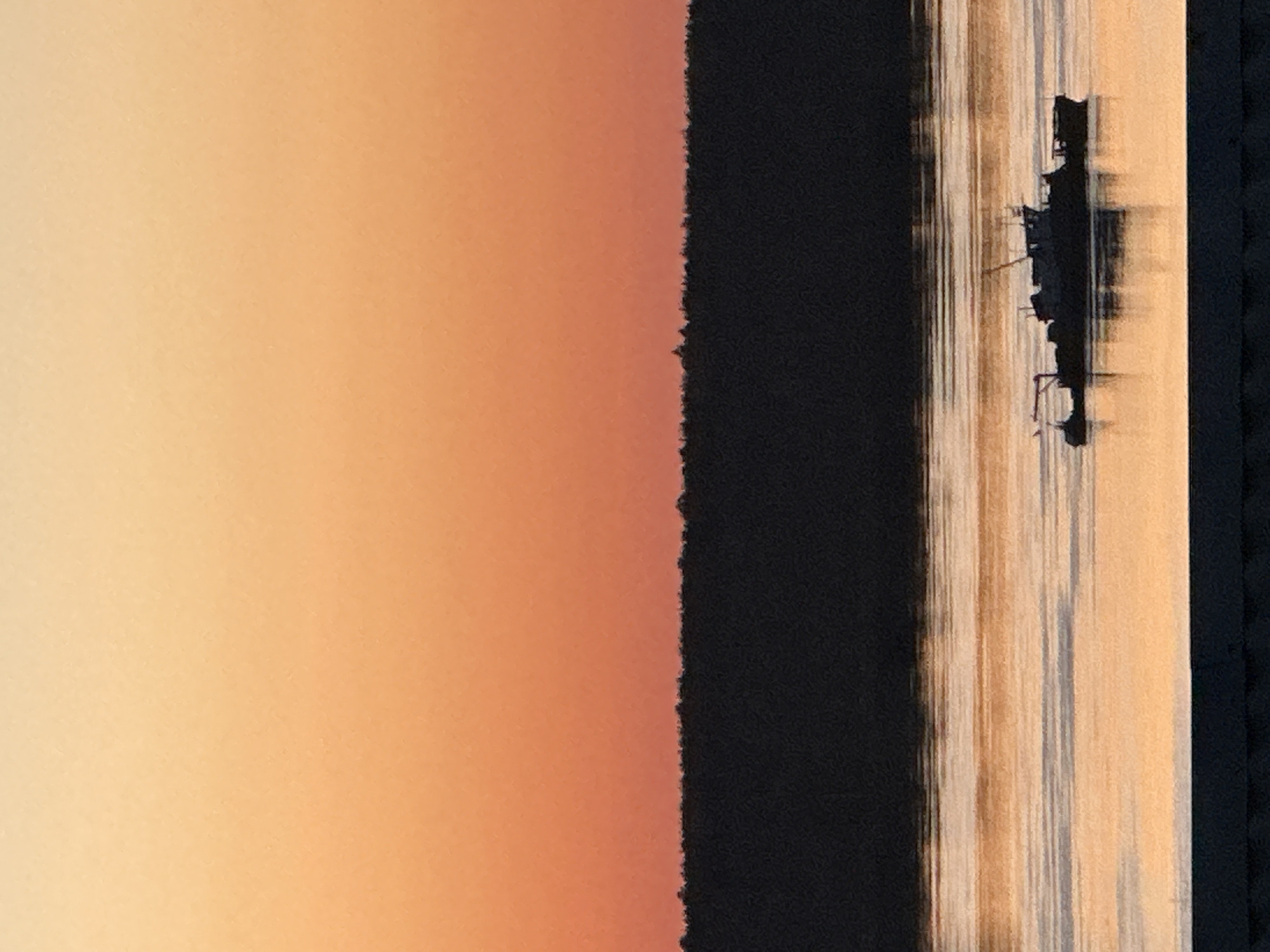 The image shows a vertical composition with a warm gradient background transitioning from light to dark orange on the left. A dark, textured vertical band separates the background from a lighter area with vertical striations that suggest water reflections. A dark, silhouetted structure resembling a boat or platform is visible within the striated section. The right edge of the image is bordered by a dark, vertical band. The image may be a photograph of the sunrise over the pier, in the city of Chaikovsky, Russia.