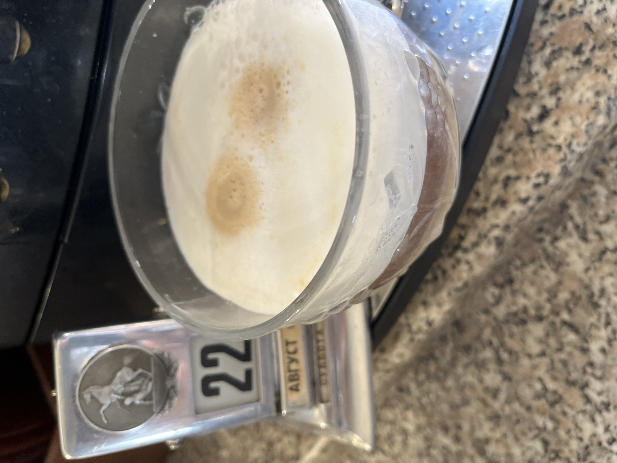 An eye-level, close-up shot shows a clear glass mug filled with a frothy, white-topped beverage, likely a coffee drink with milk or cream, sitting on a metallic surface. Two darker, circular spots of crema are visible on the surface of the foam. To the left of the mug, a silver desk calendar displays the number '22' prominently. Below the number, the word "АВГУСТ" (August) is visible in Cyrillic letters, followed by "СУББОТА" (Saturday). The background consists of a dark appliance, likely a coffee machine, and a speckled granite countertop. The scene is lit from above, casting soft shadows. No individuals are present.