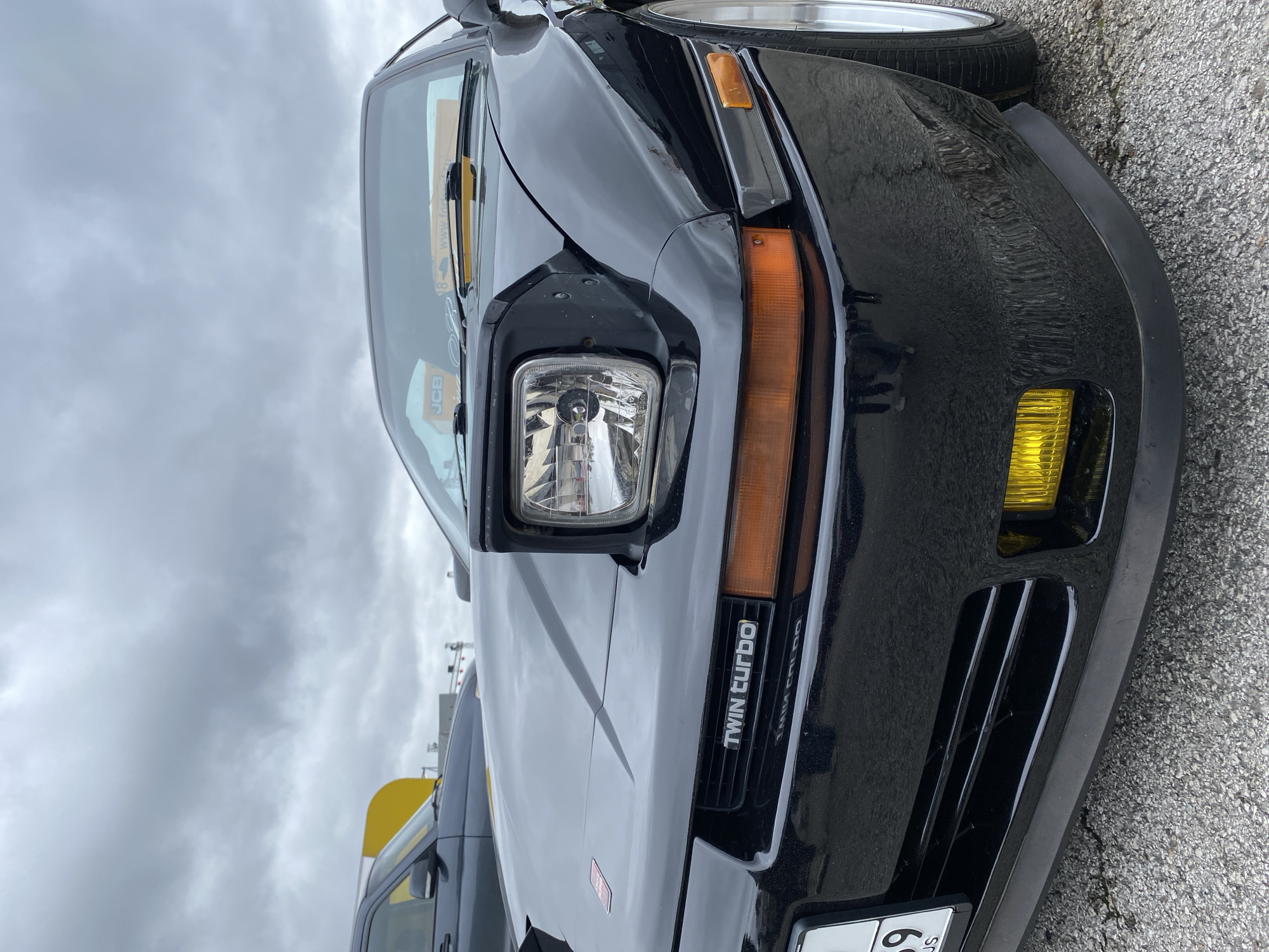 The image shows a close-up, low-angle view of the front of a black sports car, identified by the "TWIN TURBO" emblem on the grille. The car's pop-up headlight is raised, revealing a clear lens. Amber turn signals are visible below the headlight. Yellow fog lights are mounted in the lower bumper. The car is parked on a grey, asphalt surface. In the background, other vehicles and a yellow object are partially visible. The sky is overcast. Based on the license plate visible on the car which reads "274 59 RUS", the location is Russia, specifically the Perm Krai region.