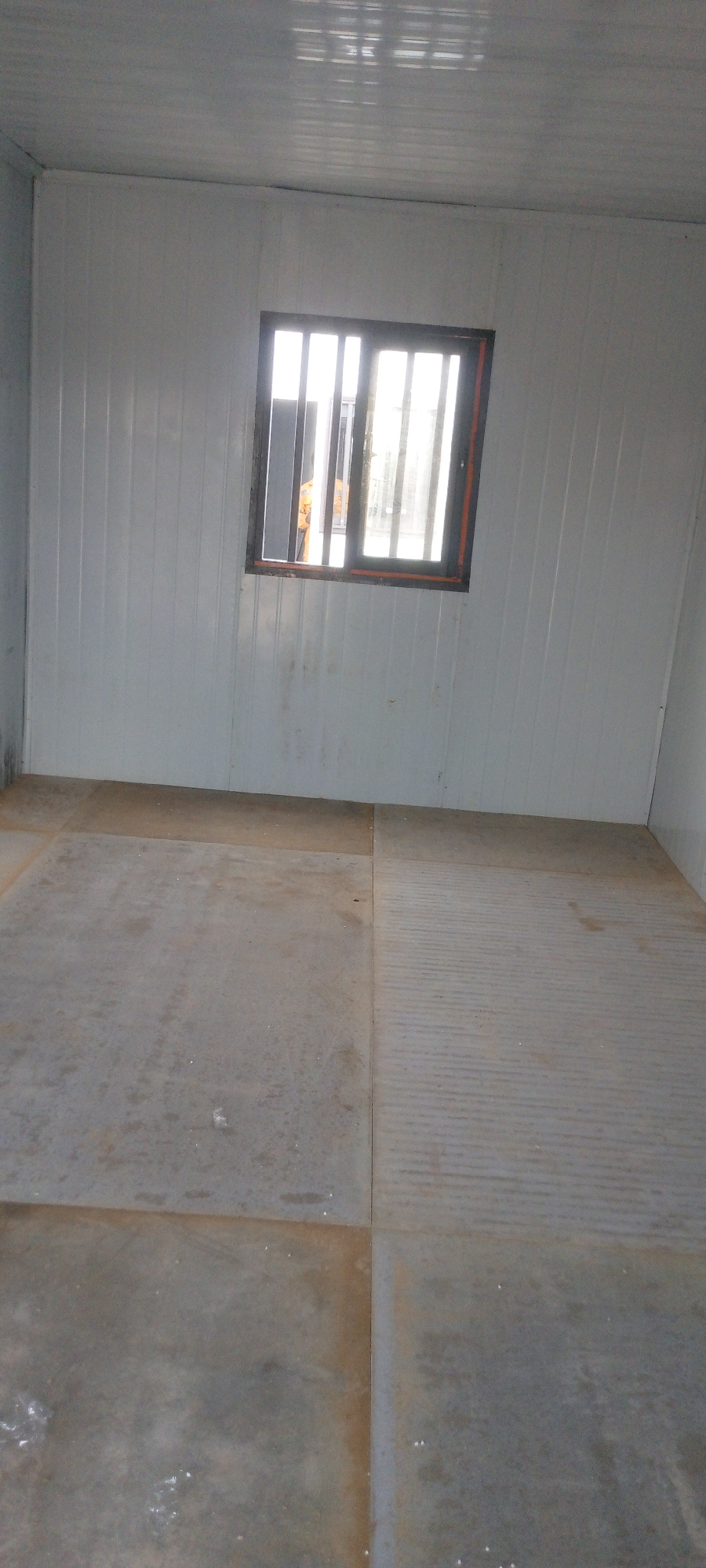 The image shows the interior of a room constructed with white corrugated metal panels on the walls and ceiling. A small, barred window is visible on the wall, with sunlight streaming through. The floor appears to be made of large, grey concrete tiles, with some sections exhibiting a ribbed texture. The room is sparsely furnished, with only the flooring and walls visible. Based on the location information provided, this scene is in Indo, Nigeria. There are no people or specific objects of note within the room, apart from the structural elements and the window. The overall impression is of a simple, utilitarian space, possibly a prefabricated structure or a basic utility room.