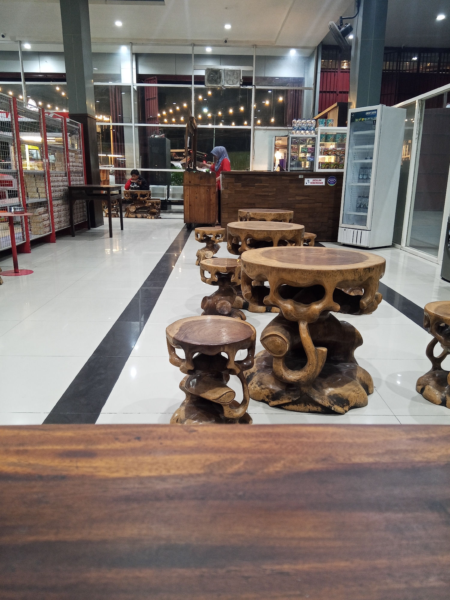 The image depicts the interior of what appears to be a cafe or restaurant located in Hasanuddin, Indonesia. In the foreground, a wooden table is partially visible, with its textured surface taking up the bottom portion of the frame. Beyond this, a row of intricately carved wooden stools and low tables are arranged on a white tiled floor, accented by a black stripe running down the center. The stools and tables have a rustic, natural wood aesthetic, with some featuring abstract carved bases resembling roots or natural forms.
In the mid-ground, a woman wearing a blue headscarf and a red shirt stands behind a wooden counter, seemingly attending to a customer or preparing something. A young boy in a red shirt sits at one of the wooden tables, looking down at something on the table. Further back, a large glass-fronted refrigerator is visible, stocked with beverages. Behind the counter, shelves are stocked with various items. The background features a wall with large windows, offering a view of the exterior, where more lighting and structures are dimly visible. The overall ambiance suggests a casual, possibly artistic, establishment.