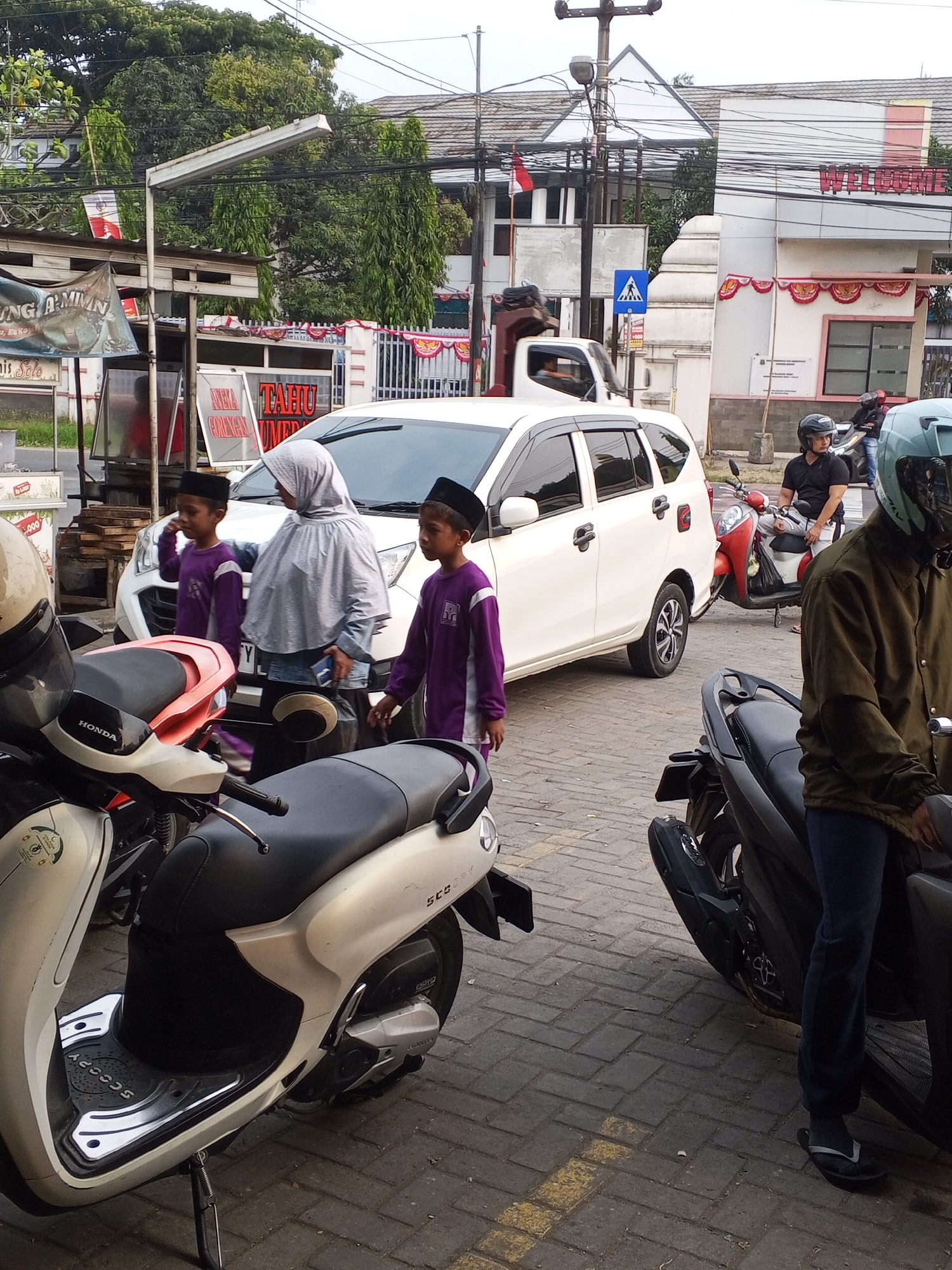 In the city of Ciruas, Indonesia, a street scene unfolds with a white multi-purpose vehicle parked on a brick-patterned paved road. Two young boys, wearing purple long-sleeved shirts and black peci caps, stand near the car. One boy is looking towards the car, while the other stands facing left. A woman wearing a grey hijab and a light-colored top is standing between the boys, looking towards the car and holding a dark object in her hand.
Several motorcycles are visible. A white and orange scooter is parked in the foreground on the left, with its handlebars turned left. To the right of the white MPV, a red scooter is parked, with a man in a black shirt and white pants wearing a helmet sitting on it. Further to the right, another motorcycle is parked, and a person wearing a green helmet and a brown jacket is partially visible, sitting on it.
In the background, buildings with white and red decorative elements are visible, along with trees, power lines, and streetlights. A sign with the word "WELCOME" in red letters is mounted on one of the buildings. A pedestrian crossing sign is also visible. Faded yellow lines mark the road.