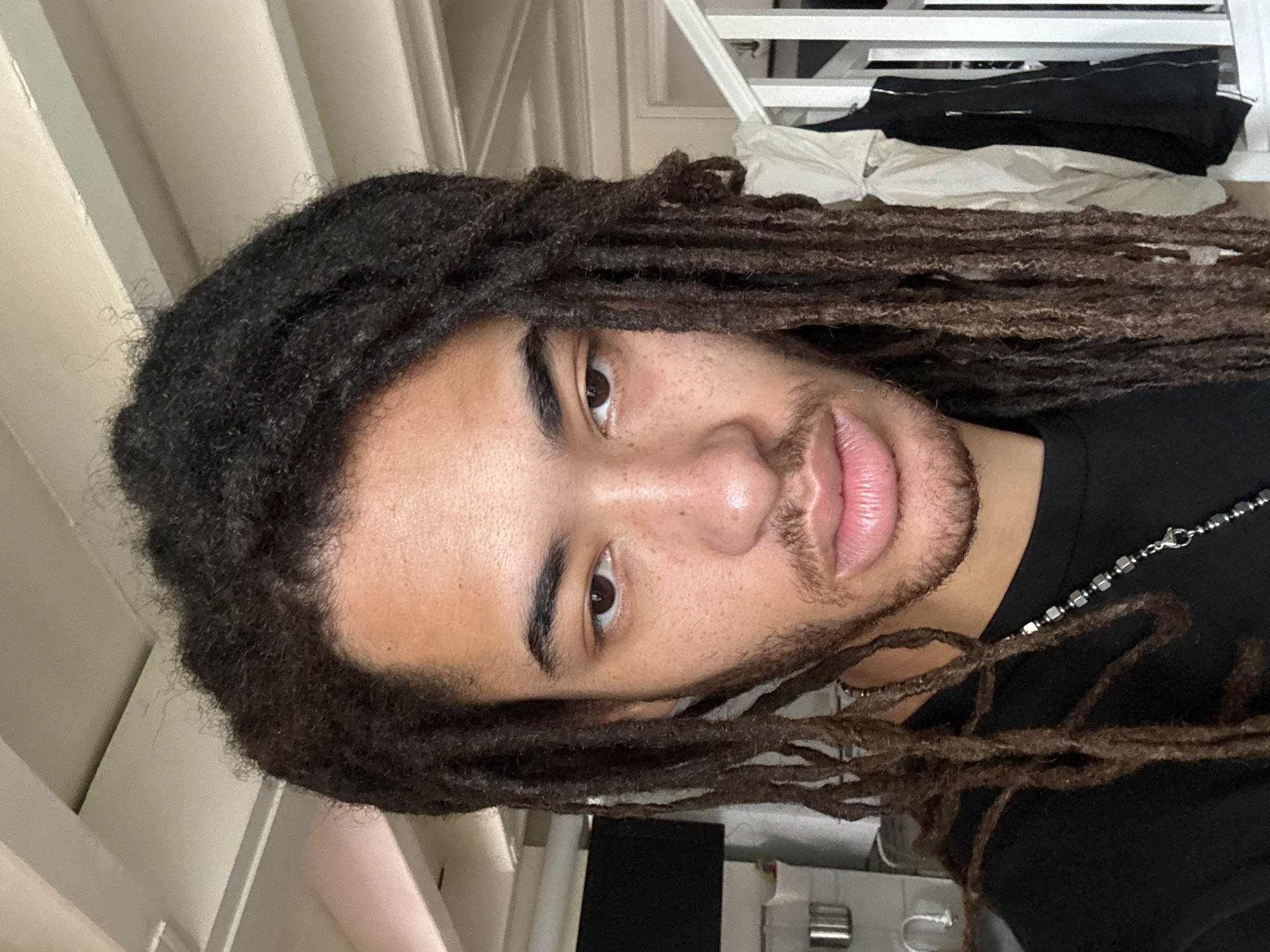 A medium shot shows a light-skinned man with locs wearing a black shirt and a necklace. He is indoors in a room with white walls and a white set of blinds. The location of Paris, France cannot be determined from the image provided.