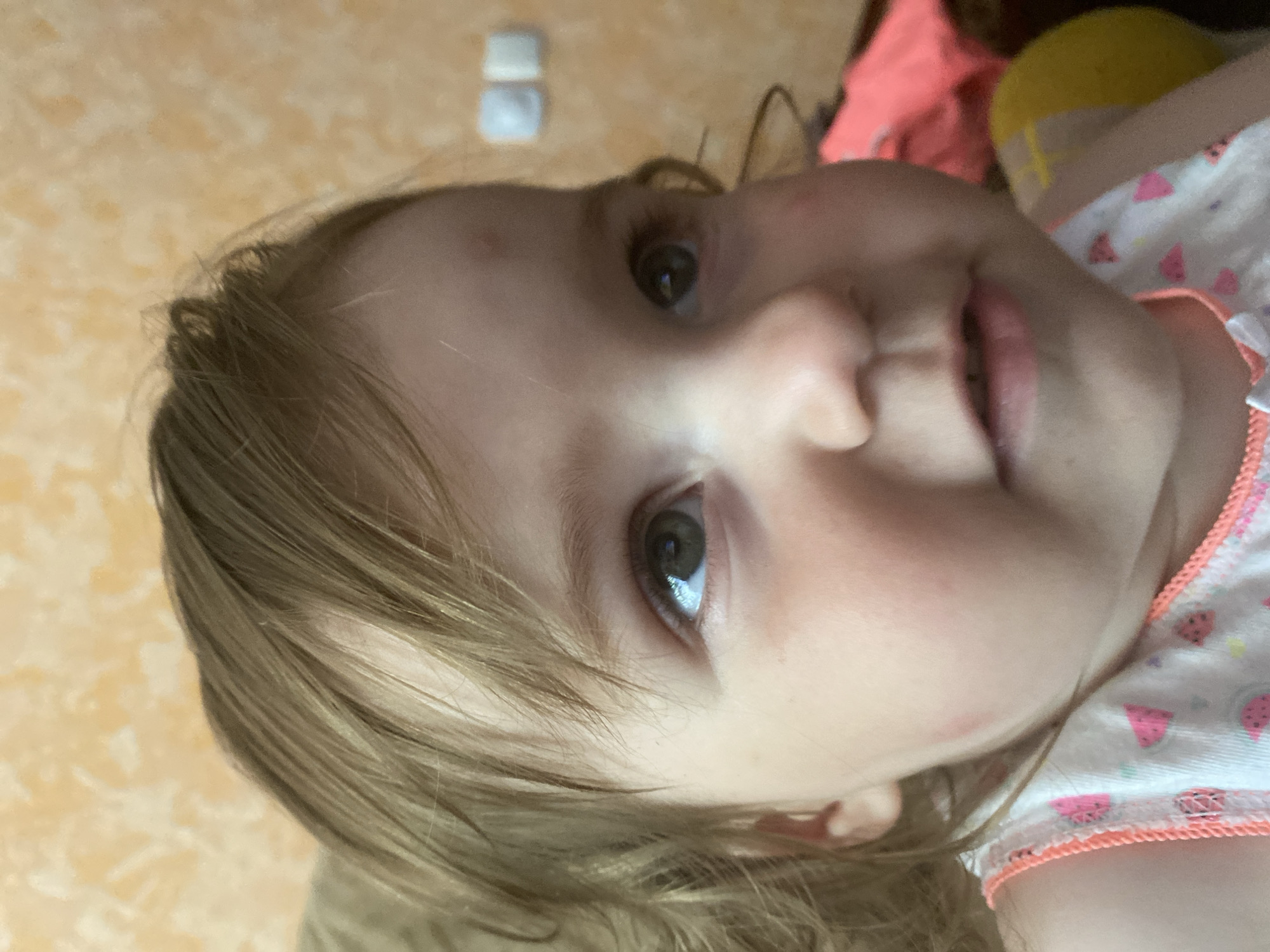 This is a close-up, low-angle photograph of a young child's face, with the camera oriented vertically. The child, appearing to be a toddler, has light brown hair that falls across their forehead and cheek. Their eyes are open and looking slightly to the left of the frame. The child's skin is fair, and their cheeks are rounded. A hint of pink is visible on their left cheek, possibly a blush or a mark.
The child is wearing a white top with a pattern of watermelons and other fruits, trimmed with a coral-colored edge. The background is blurred and appears to be a wall with an orange-yellow textured pattern, possibly wallpaper or painted plaster. In the upper left portion of the image, two small, rectangular white objects are visible, their purpose unclear.
The lighting suggests it is daytime, possibly indoors, with soft light illuminating the child's face. The overall impression is one of innocence and a candid moment captured. There is no discernible text in the image. The activity appears to be the child simply looking at the camera or something just out of frame. The emotion conveyed is neutral to curious.