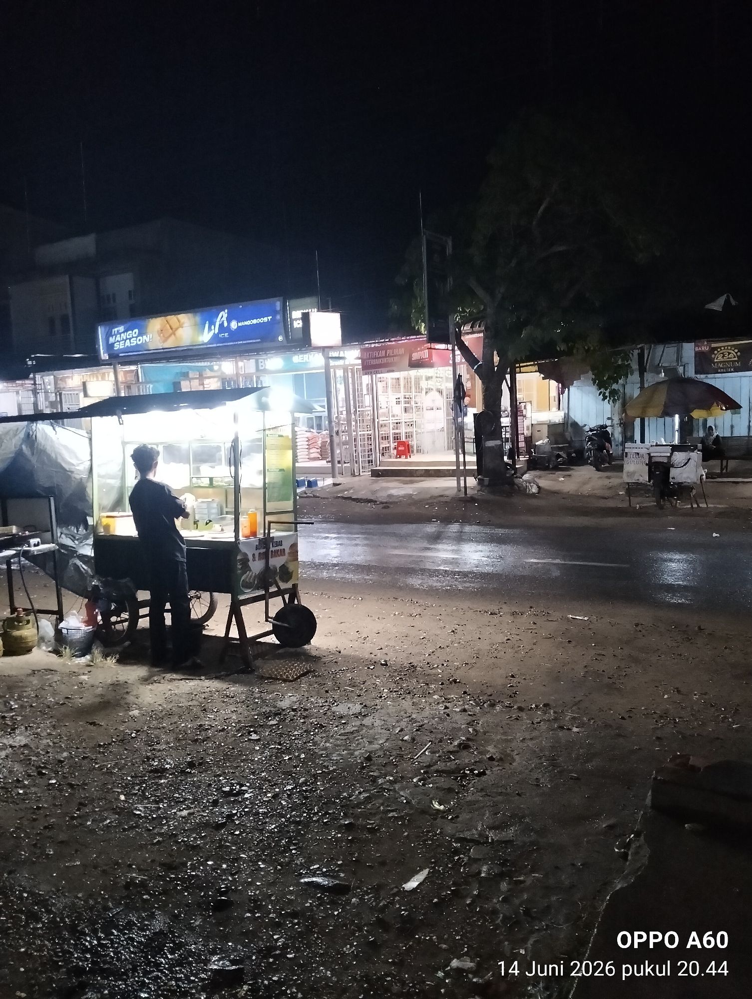 A nighttime street scene in Payadua, Indonesia depicts a food cart vendor at work. The vendor, a person in dark clothing, stands behind a brightly lit food cart, appearing to be preparing food. The cart displays items like "BURGER KEBAB & BAKAR" and a drink bottle. In the background, various shops and stalls are visible, including one with a "MANGOBOOST ICE" sign. The street surface is wet and appears to be paved with asphalt and gravel, with puddles reflecting the lights. A large tree stands near the shops. The overall atmosphere suggests a local marketplace or street with ongoing commercial activity at night.