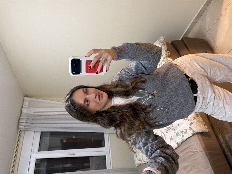 A woman in a grey sweater and white pants poses for a selfie. Her phone has a white and red patterned case. She has long brown hair and is wearing a necklace. Behind her, a window is visible with white curtains. She is sitting on a beige sofa with a patterned pillow. The walls are painted a neutral color.