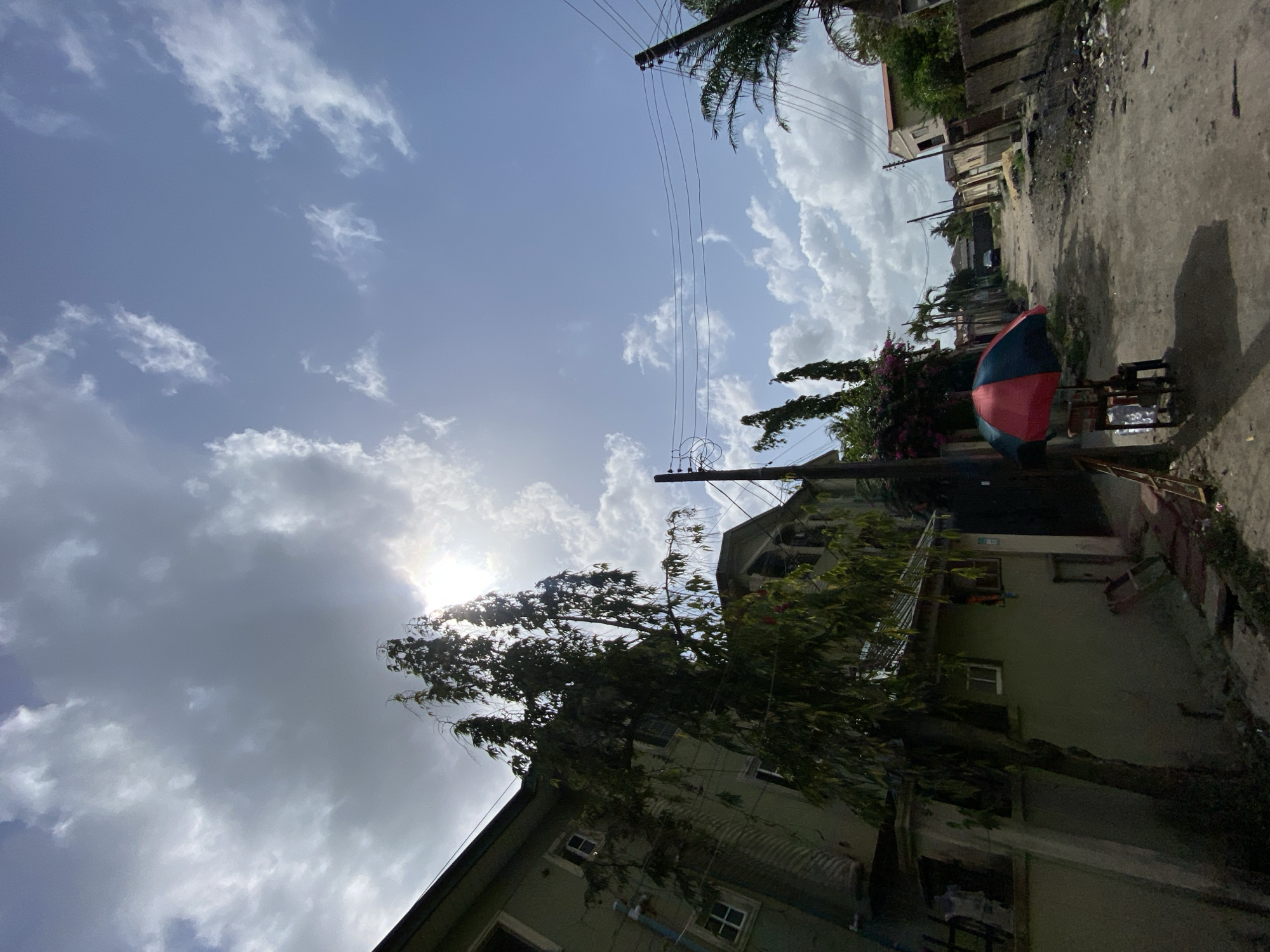 The image captures an outdoor scene, likely in a residential or semi-urban area, under a bright, partly cloudy sky. The perspective is slightly upward-angled, emphasizing the sky and tree canopy.
The sky, occupying a significant portion of the frame, is a clear blue with scattered white cumulus clouds. The sun is intensely bright, peeking from behind clouds in the upper left quadrant, creating a strong glare and lens flare. This indicates a sunny day, likely mid-morning to early afternoon.
In the lower left, a large tree with abundant green foliage stands prominently, its branches reaching towards the center. Below it, part of a light-colored two-story building with visible windows and a roofline is seen. To the right of the tree, an unpaved, dusty road stretches from the mid-ground towards the background.
Along the right side of the road, a street vendor's stall is visible, characterized by a distinctive red and blue (or black) striped umbrella providing shade. Underneath the umbrella, items resembling stacked plastic containers or bottles are arranged on a low stand, suggesting products for sale. Further along the road in the background, more buildings of varying sizes and designs, interspersed with green trees and bushes, line the pathway.
Utility poles with multiple power lines crisscross the upper and middle sections of the image, running diagonally across the sky and through the tree canopy. Palm fronds are also visible at the very top right. There are no people visible, and no discernible text. The scene depicts a common street view, indicative of the provided context of Ado, Nigeria.