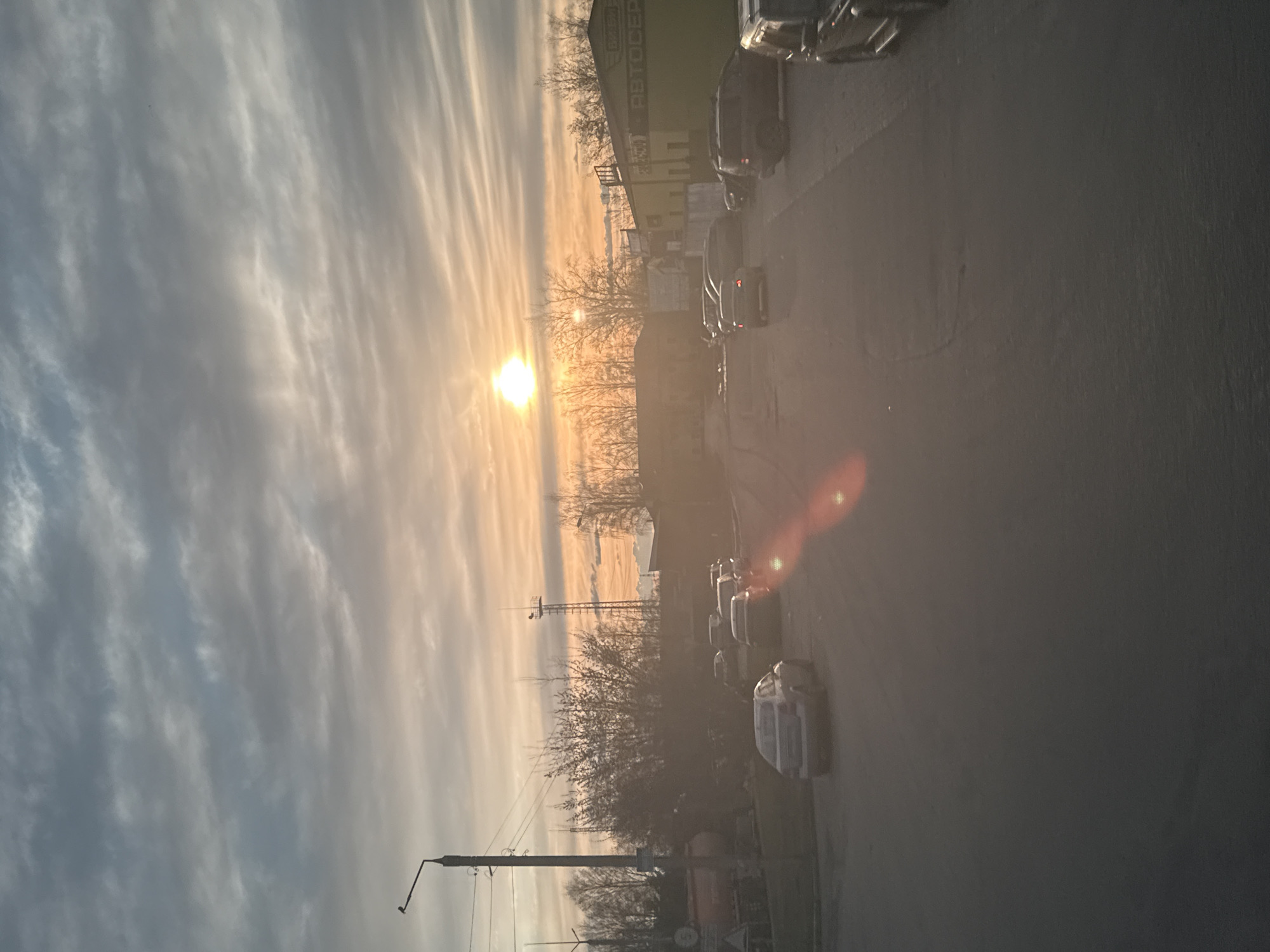 The image depicts an urban scene during a sunrise or sunset. The sky is filled with streaky clouds, illuminated by the sun positioned near the horizon. A street lined with parked cars is visible on one side. Buildings are present, including one with a sign in Russian, "АВТОСЕРВИС" (Avtoservis), suggesting a car service or repair shop. Bare trees are interspersed throughout the scene. Utility poles with wires are noticeable in the foreground. A communication tower is visible on the horizon. The location appears to be in Dmitrov, Russia, based on the Russian signage.