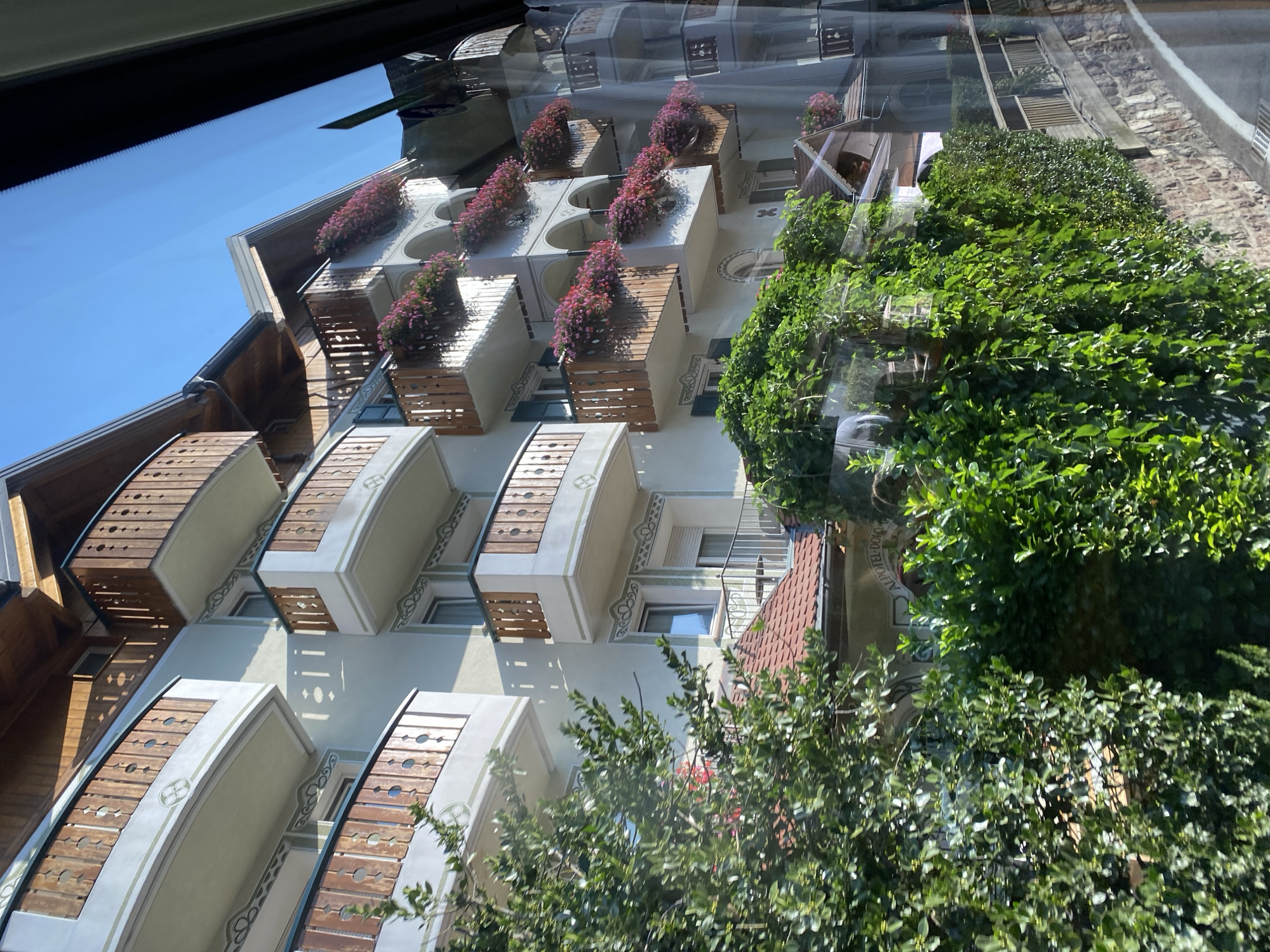 The image shows a detailed view of a hotel exterior in Molveno, Italy, under a clear blue sky. The hotel building is light-colored with ornate detailing on its balconies and facade. Wooden balconies with decorative railings extend outwards from each floor, adorned with abundant pink and red flowers in planters. The building features multiple stories, with arched windows and shutters on some levels. Lush green foliage and trees partially obscure the lower sections of the hotel and the surrounding grounds. The architectural style suggests a traditional alpine or European design, possibly reflecting the mountainous region of Molveno. The image is captured at an angle, giving a sense of depth and perspective to the structure.