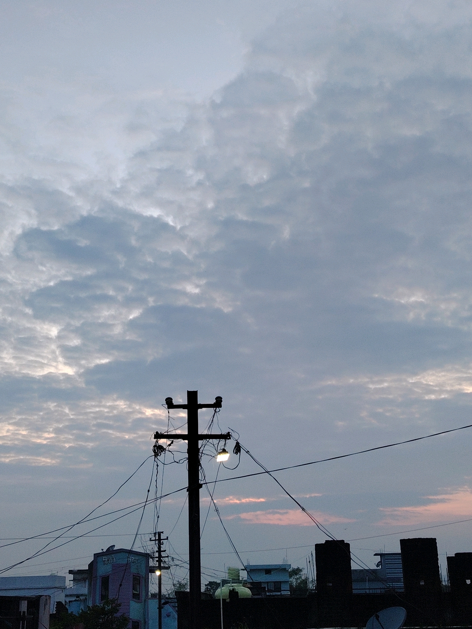 This is a vertical, medium shot of a sky filled with scattered clouds, viewed from a rooftop in Sihora, India. In the foreground, a silhouetted telephone pole with many wires and two lights stands prominently. To the left of the pole, a multi-colored building with text in Hindi is visible. The middle ground features a line of dark silhouettes of buildings and water tanks. The background is dominated by the sky, which exhibits subtle hues of pink and orange near the horizon, suggesting either sunrise or sunset. The overall atmosphere is serene and muted.