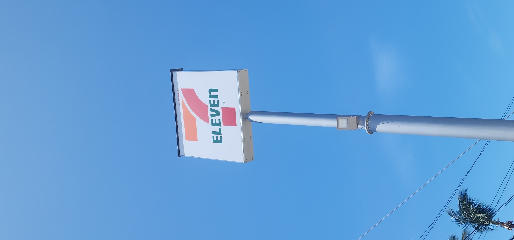 A tall, grey metal pole stands against a bright blue sky, supporting a white rectangular sign with the "7-Eleven" logo. The logo features a large orange and pink "7" positioned above the green text "ELEVEN" with a small red square beneath it. A white LED light fixture is attached to the pole, about a third of the way up. In the lower left corner, the fronds of a palm tree are visible, swaying in the wind, along with some dark electrical wires. The scene appears to be outdoors, under a clear sky, possibly in a tropical location like Ko Samui District, Thailand.