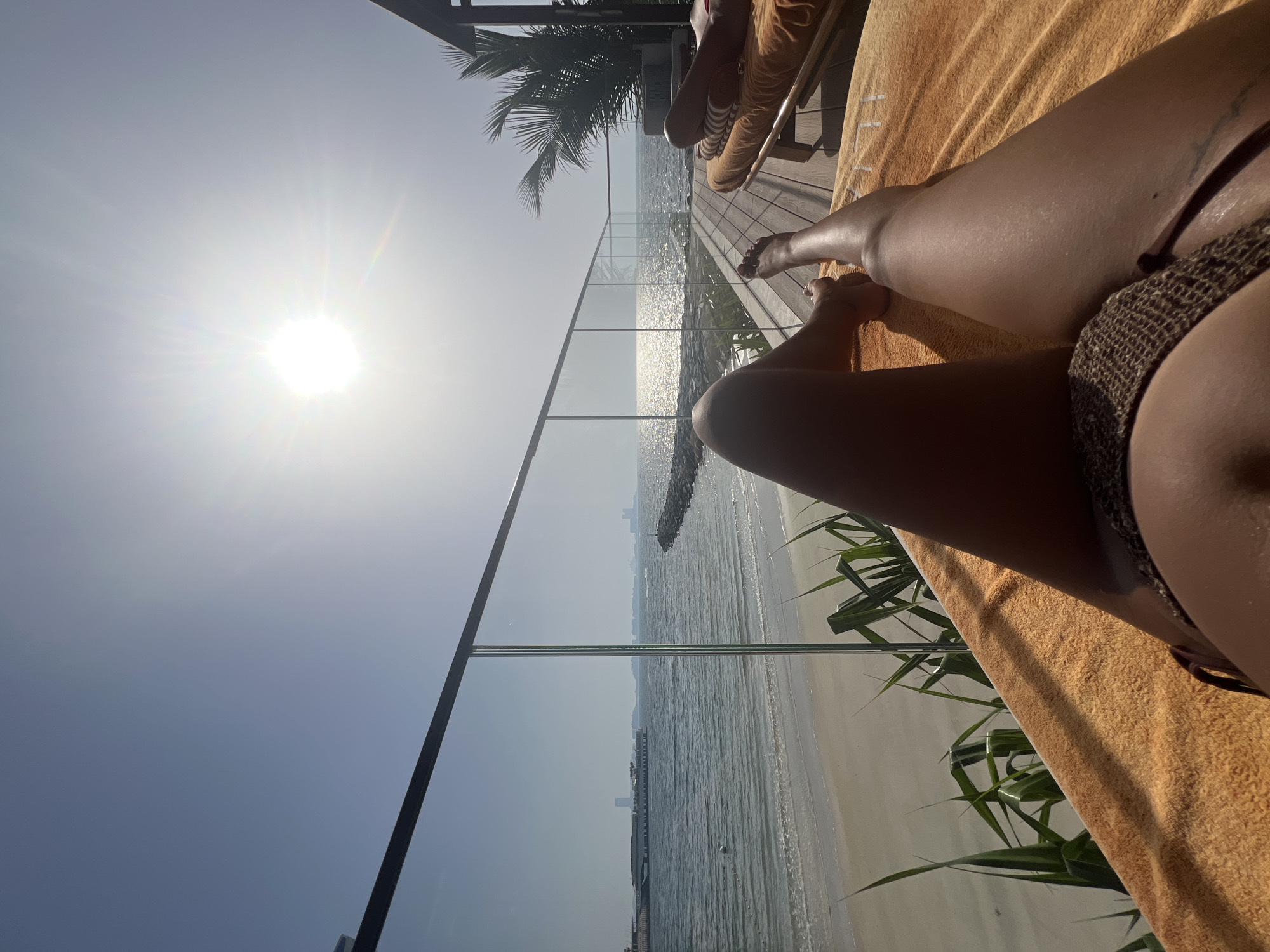 The image captures a sun-soaked scene on what appears to be a deck or balcony overlooking a beach in Dubai, United Arab Emirates. A person is reclining on an orange towel, clad in what seems to be a bikini or swimsuit. Their tanned legs are prominently featured in the foreground. The background features a glass railing that separates the deck from the beach, where the ocean stretches out to the horizon. In the distance, buildings and structures are visible, hinting at Dubai's urban skyline. To the left, a palm tree adds to the tropical ambience. The sun shines brightly, casting a glare over the scene.