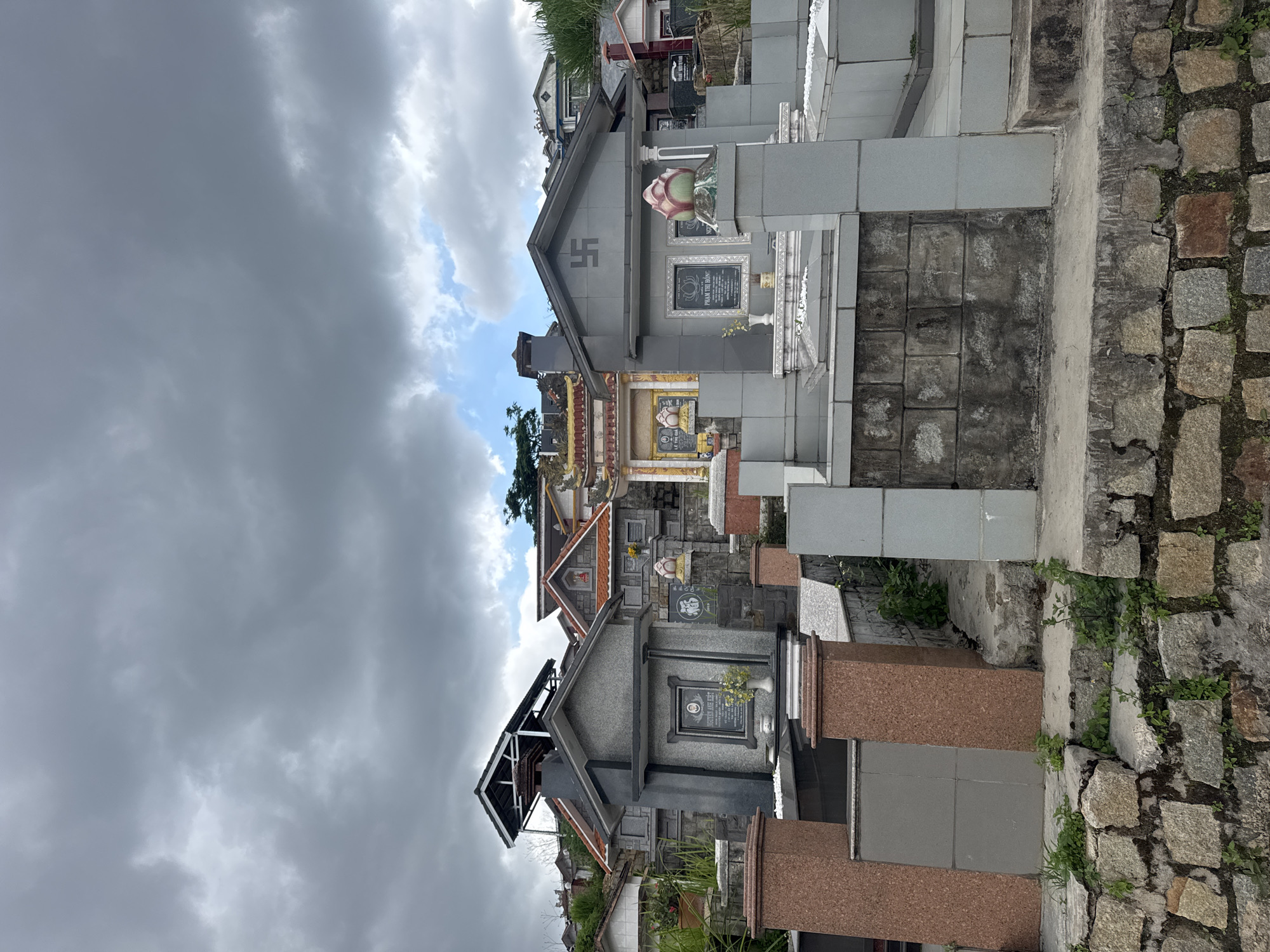 This photograph captures a cemetery in Lạc Dương, Vietnam. The tombs are densely packed and vary in architectural style, but are predominantly made of gray and brown stone and tile. Some structures are adorned with religious symbols, including a swastika (a symbol which has different meanings depending on location). The sky above is overcast with heavy, dark gray clouds, partially obscuring patches of blue sky. Vegetation grows sporadically around the stone structures and steps. The photograph is taken at a low angle, looking upwards toward the sky.