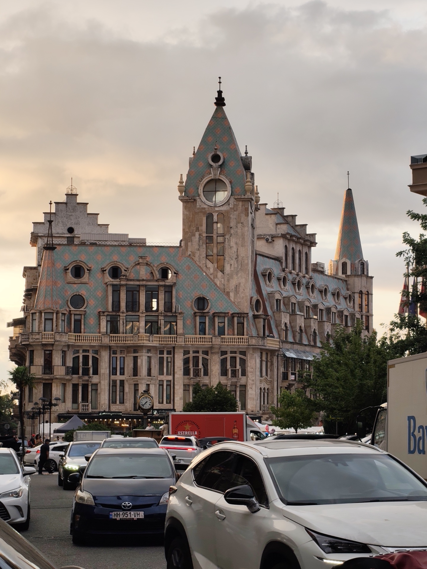 The image depicts a view of the Batumi Astronomical clock tower and the surrounding street scene in Batumi, Georgia, during the golden hour. The clock tower, with its distinctive pointed roof adorned with green and orange tiles and a large circular clock face, is the focal point. The building exhibits intricate architectural details, including multiple windows, balconies, and decorative elements on its facade.
The street is filled with traffic, featuring several cars of various models and colors, including a prominent white Lexus in the foreground, a dark blue Toyota Prius, and other vehicles. People are visible on the sidewalks and crossing the street, though their faces are not clearly discernible. Red trucks and a white delivery truck are also present on the road, suggesting commercial activity.
The background shows a mix of buildings, trees, and a cloudy sky tinged with the warm hues of sunset. Streetlights and awnings are also visible, contributing to the urban setting. The overall atmosphere is that of a bustling city street at dusk, with the architectural splendor of the clock tower standing out against the urban landscape and the soft evening light.