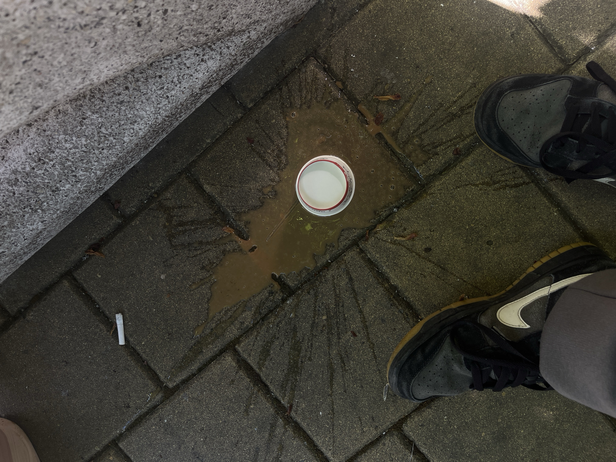The scene appears to be an outdoor public area, possibly a sidewalk or patio, likely in Karnobat, Bulgaria. It's constructed of interlocking concrete pavers. A white paper cup with a red rim rests on the pavers, surrounded by a dark, murky liquid. The liquid appears to have spilled, creating a radial pattern outward from the cup. To the left of the cup, a single, discarded cigarette lies on the ground. Two shoes of a person are present; the shoe brand is Nike, specifically of the "Dunk" silhouette with a white swoosh. The shoes appear worn, likely showing signs of regular use. The shoe to the right is resting on the same surface as the cup and liquid. Part of a concrete structure, possibly a low wall or building foundation, is visible in the upper left corner. The pavers are stained, and there are small organic debris scattered around.