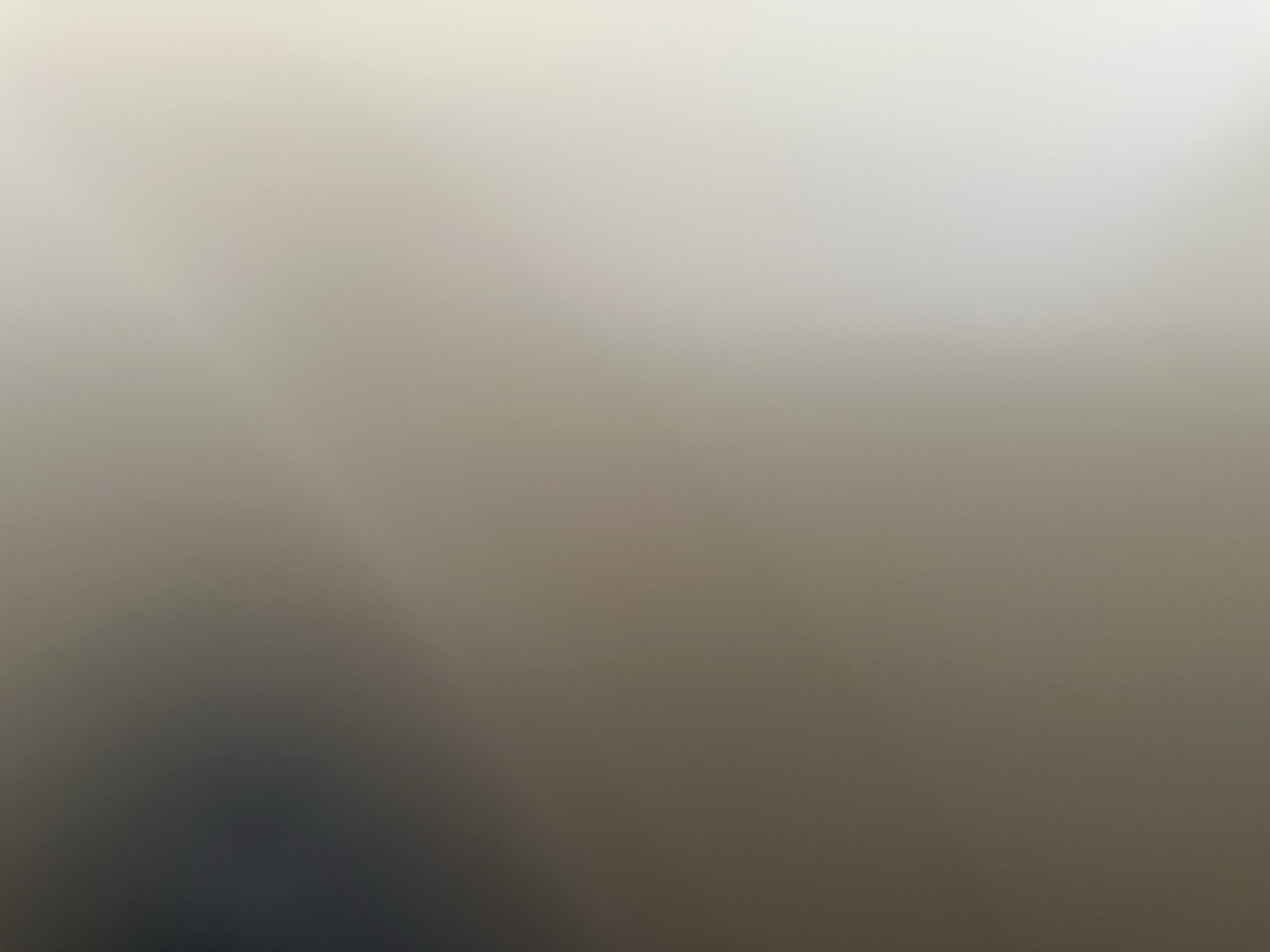The image is a blurred, abstract shot with no discernible subjects, objects, or scene. It appears to be an interior shot due to the diffused lighting, which suggests artificial light rather than natural sunlight. The color palette is muted, dominated by shades of beige, grey, and a darker, shadowy area in the bottom left corner. There are faint diagonal lines of light, possibly indicating light sources or reflections. The overall impression is one of ambiguity and lack of detail, making it impossible to determine any specific activity, time of day, weather, location cues, or emotions. No text is visible in the image.