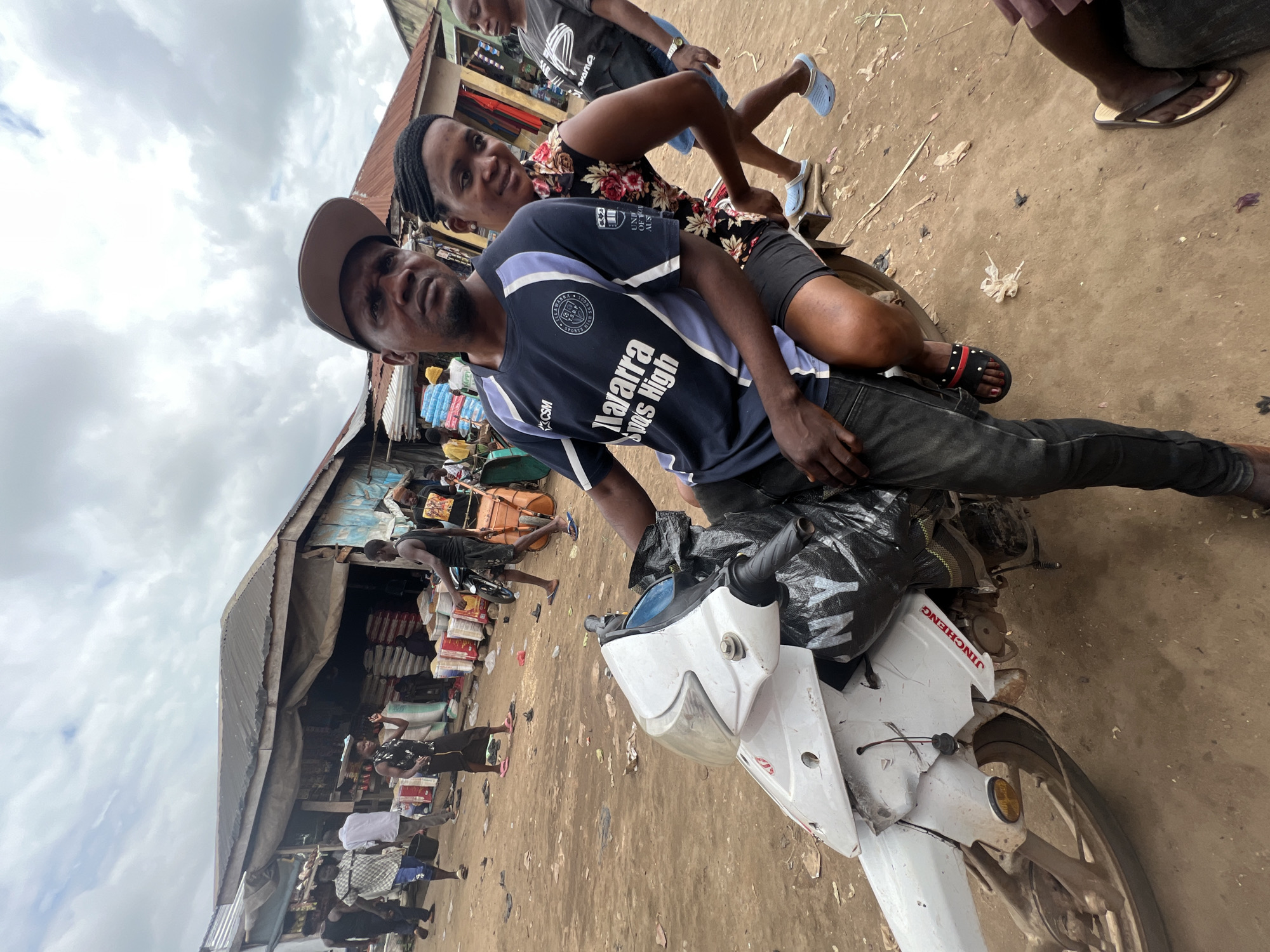 A man and a woman are seated on a white motorcycle in a marketplace in Oko Amakom, Nigeria. The man, wearing a cap and a dark blue t-shirt with white and light blue accents, is in the driver's seat. The woman, with braided hair, is seated behind him, wearing a floral print top. A black plastic bag is tied to the front of the motorcycle. In the background, market stalls are visible with people walking and interacting. The ground is dirt, and the sky is overcast. The motorcycle appears to have some damage to its front fairing, with red lettering that reads "JINCHENG" visible on the side. The overall scene depicts a busy, everyday moment in a Nigerian marketplace.
