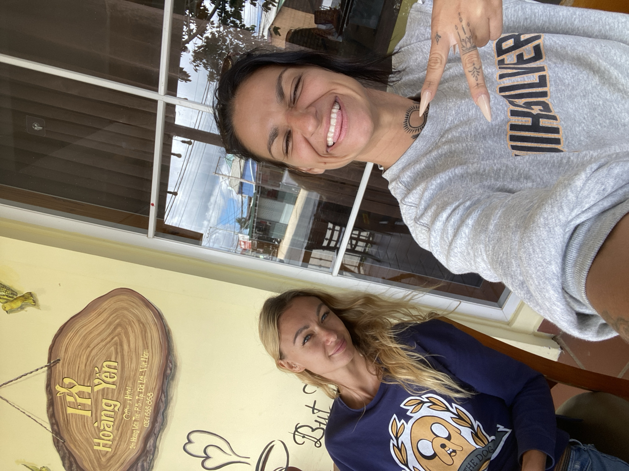 Two women are posing for a selfie in front of a window at a cafe/hotel in Da Lat, Vietnam. The cafe, named "Hoang Yen," has a wooden sign with Vietnamese writing and decorative elements. The woman on the left has blonde hair and is wearing a dark blue sweatshirt with a cartoon dog graphic. The woman on the right has dark hair and is wearing a grey sweatshirt. She is making a peace sign with her fingers and has a sun tattoo on her neck. The background shows a street scene with buildings and trees through the window. The environment appears to be outdoors during the daytime.