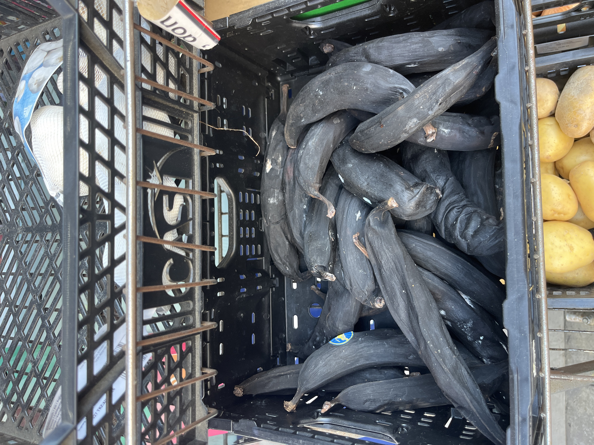 A close-up, eye-level shot shows a black plastic crate filled with very ripe, dark-skinned plantains. The plantains are tightly packed, with some showing signs of bruising and splitting. Some also have small blue stickers on them. Above the crate of plantains, a metal rack is visible, containing another black plastic crate with what appears to be packaging or bags. The background is slightly out of focus, showing hints of green and other colors, suggesting an outdoor market or produce stand. The scene is devoid of people, and no specific location in Geneva, Switzerland, can be identified from the image.