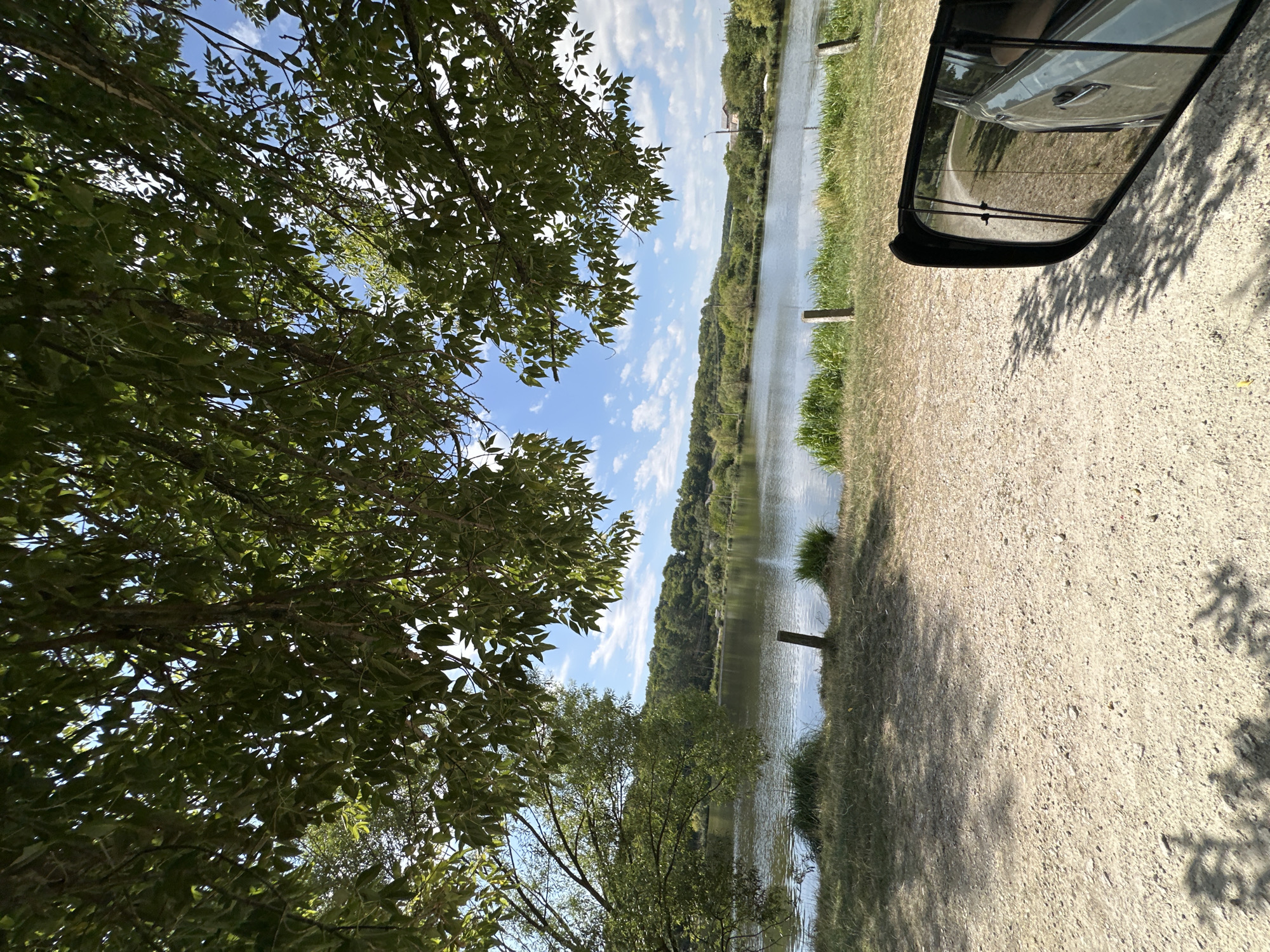 The image depicts a serene lakeside scene on a sunny day, framed by the lush green foliage of overhanging trees. The foreground is a gravelly path, casting dappled shadows from the leaves above. To the right, a car's side-view mirror reflects the surrounding environment, showing a glimpse of the car's exterior and the road behind. Beyond the path, a strip of dry grass and some taller green reeds lead to the calm surface of a lake. The water reflects the bright blue sky dotted with fluffy white clouds. Across the lake, a rolling hillside is covered in a dense forest. In the distance, a few buildings are visible amongst the trees on the right side of the image. The location is identified as being in the city of Makiv, Ukraine. The scene is static and peaceful, suggesting a moment of stillness and natural beauty.