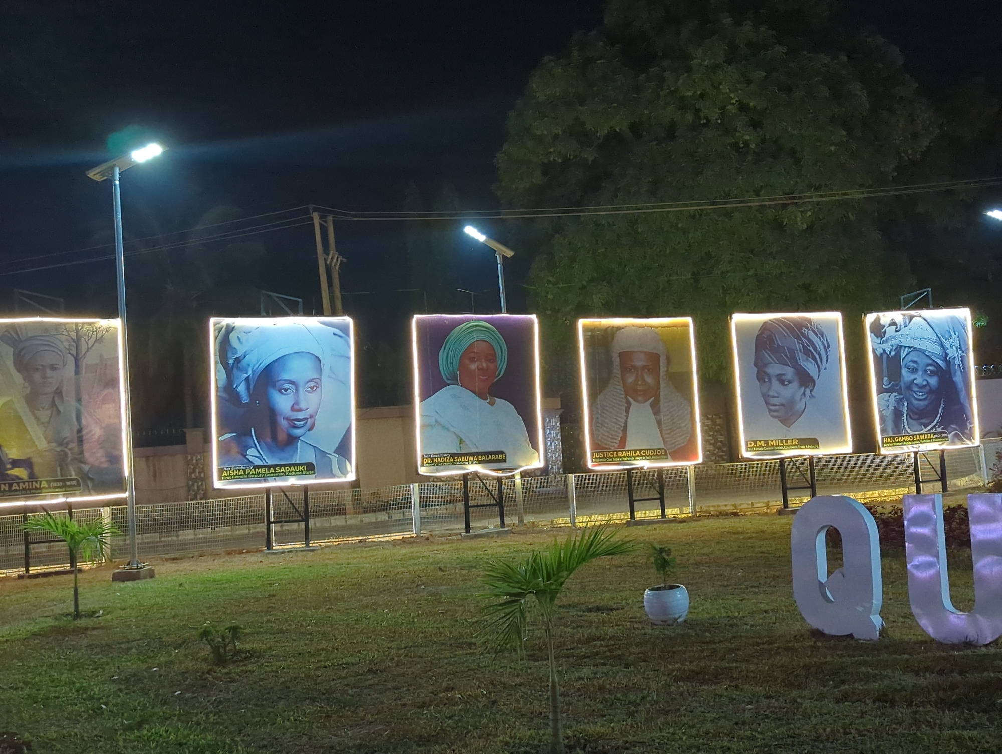 The image shows a park in Unguwar Rimi, Nigeria, at night. There are six illuminated portraits of women, arranged in a row, likely a memorial or monument. The portraits are framed and displayed on posts. The subjects include Queen Amina, Aisha Pamela Sadauki, Dr. Hadiza Sabuwa Balarabe, Justice Rahila Cudjoe, D.M. Miller, and Haj. Gambo Sawaba. The portraits include each woman's name and a brief description of her accomplishments or role. In the foreground, there is a grassy lawn with young palm trees and other small plants. The letters "QU" are present in large, possibly illuminated, characters. Overhead streetlights provide illumination. A wire fence runs behind the portraits.